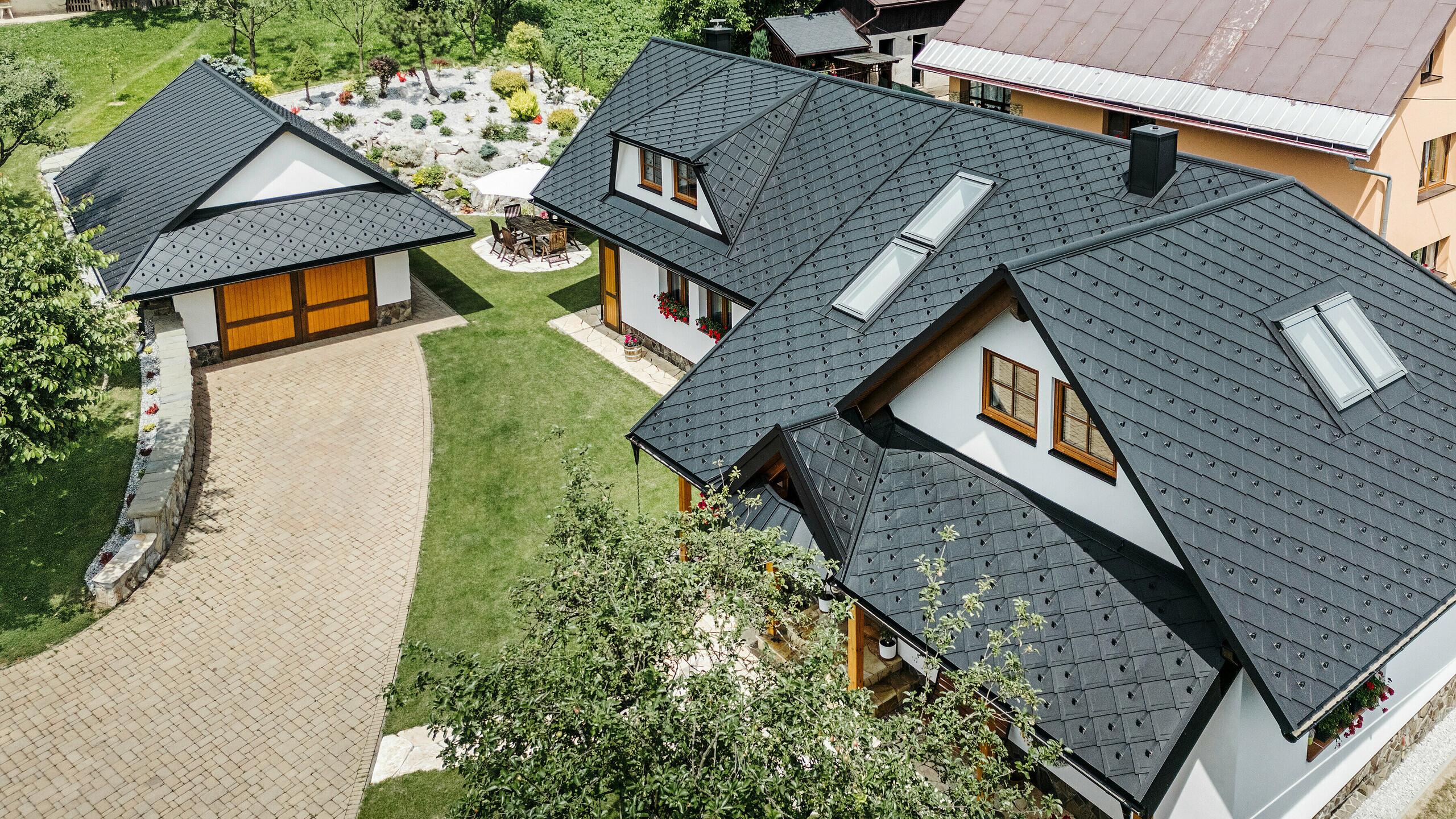 Vue aérienne d'une maison familiale à Stara Bystrica, en Slovaquie, avec un toit en losanges PREFA 29 × 29 en P.10 noir. Le toit, conçu dans un aspect de diamant uniforme, comprend des fenêtres de toit inclinées et des lucarnes trapézoïdales des deux côtés. Les décorations en bois sur les fenêtres et les compositions florales colorées confèrent à la maison un aspect invitant et harmonieux. Le jardin de devant bien entretenu et l'allée pavée complètent parfaitement l'ensemble.