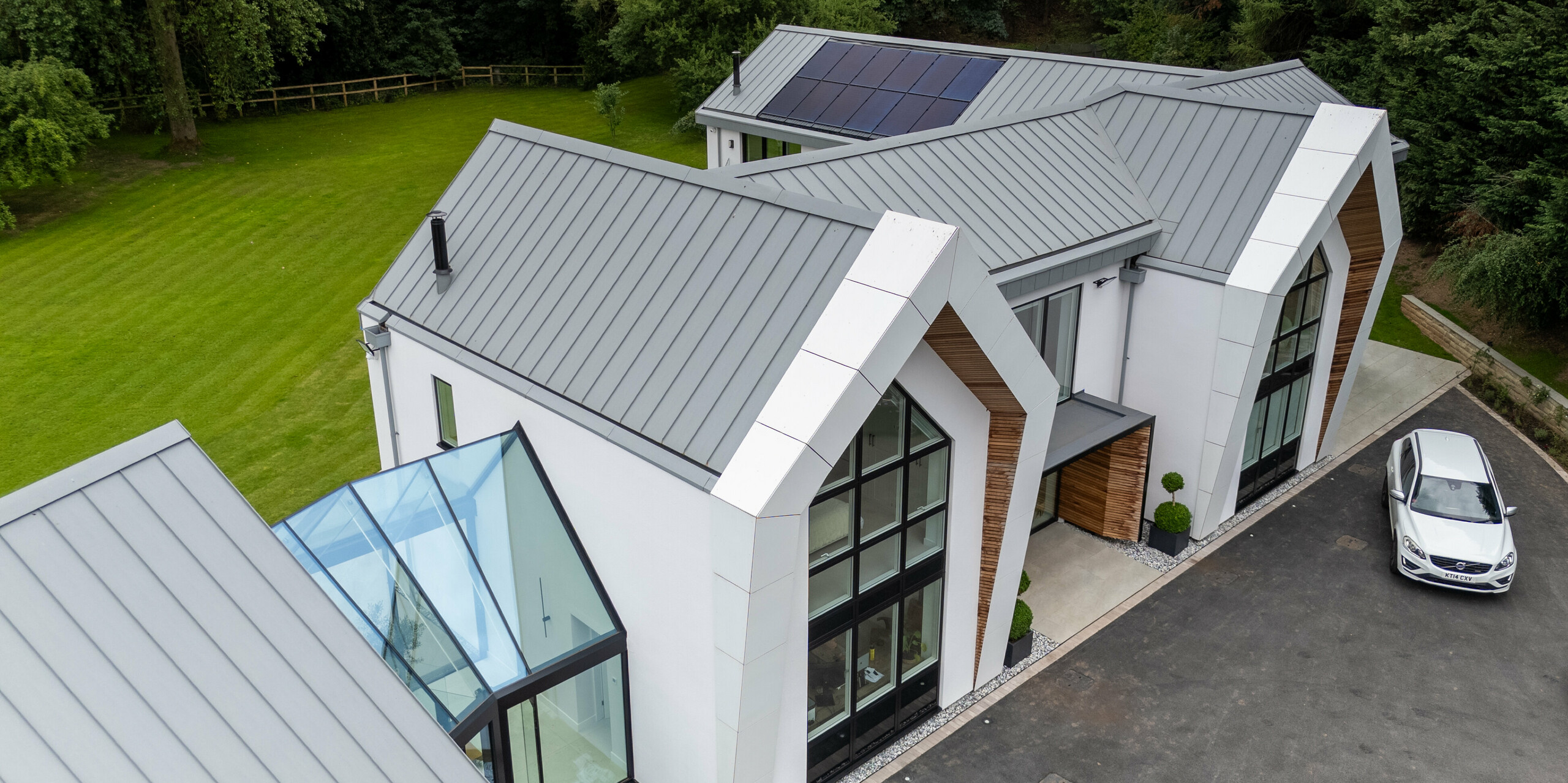 L'image présente la villa moderne de Draycott-in-the-Clay sous un angle saisissant. Son élégante toiture PREFALZ gris quartz attire immédiatement le regard. La structure à joints debout épurée confère à l'architecture une allure moderne et distinctive, et s'intègre harmonieusement au paysage verdoyant. L'image met également en valeur l'alliance réussie des surfaces vitrées et du bardage bois en façade, qui, associée à la robustesse de la toiture PREFALZ, crée une esthétique raffinée. Le système photovoltaïque installé sur le toit témoigne de l'engagement de la maison en faveur du développement durable.