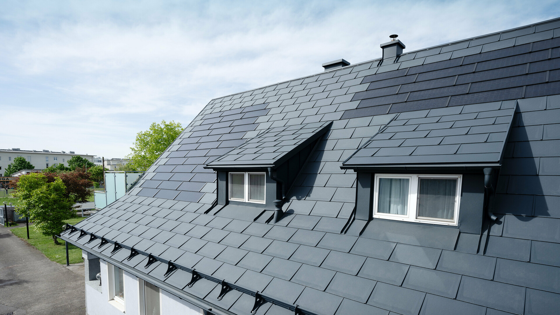 Gros plan sur un toit recouvert de grandes tuiles solaires PREFA et de tuiles PREFA R.16 en P.10 anthracite. On aperçoit deux lucarnes. Les tuiles solaires sont réparties de manière homogène sur la toiture et offrent une solution moderne et durable.