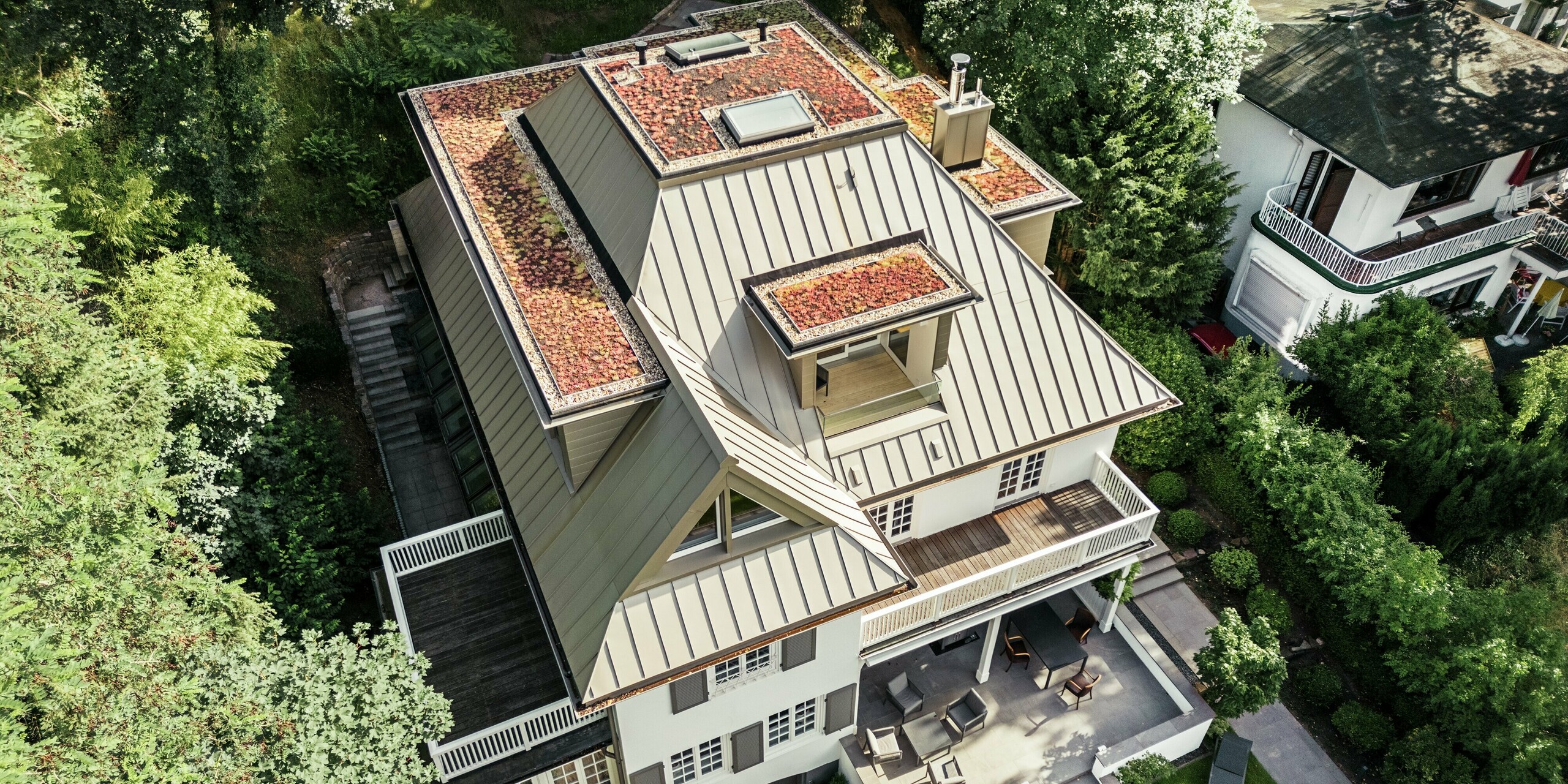 La photographie aérienne révèle la Villa Blankenese à Hambourg et son impressionnante toiture PREFALZ couleur bronze. La forme complexe de cette toiture, avec ses multiples lucarnes et ses surfaces intégrées, est mise en valeur par l'élégante technique de la pose à joints debout. Le bardage Siding PREFA couleur bronze des éléments de façade complète harmonieusement l'ensemble, créant une continuité visuelle entre la toiture et la façade. L'alliance de l'aluminium résistant et d'une teinte sophistiquée confère à l'édifice un style élégant et une résistance aux intempéries qui attirent tous les regards au cœur de son environnement verdoyant.