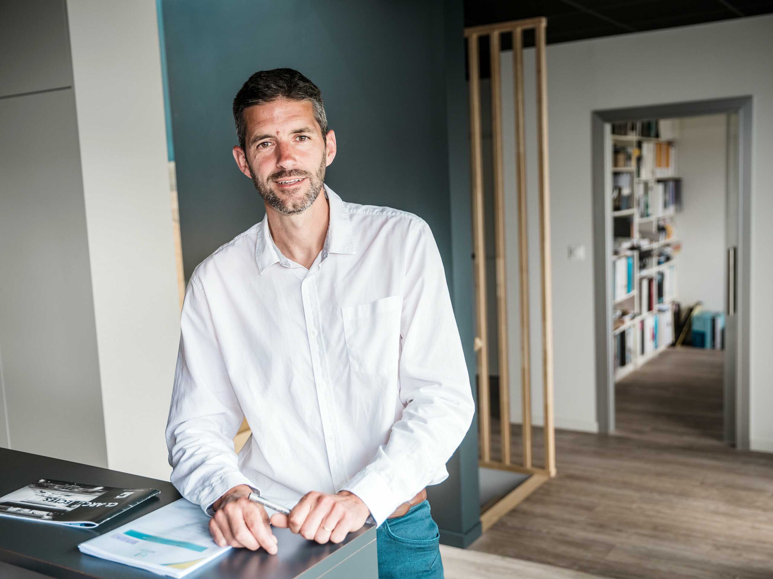 Au premier plan, l'architecte Hugues Launay, du cabinet CLARC Architectes, est positionné au centre, légèrement à gauche, et vêtu de façon formelle, comme il se doit pour un entretien professionnel. À l'arrière-plan, la façade incurvée en aluminium du Collège Léontine Dolivet, dont la surface argent métallisé crée des reflets lumineux. M. Launay regarde légèrement sur le côté ; sa posture, empreinte d'assurance et de confiance en soi, suggère un rôle de chef de projet. La scène a été photographiée en lumière naturelle douce, qui met en valeur les contours avec netteté.