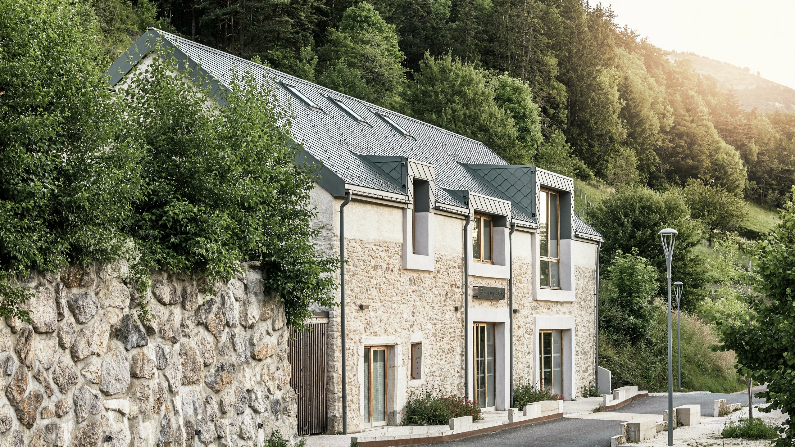 Le corps de ferme restauré "La Grange à Gaby" à Engins, France. Le bâtiment combine la maçonnerie en pierre traditionnelle avec un toit moderne en aluminium PREFA composé de losanges de toiture de 29 × 29 cm. La rénovation allie construction historique, architecture contemporaine et matériaux durables.