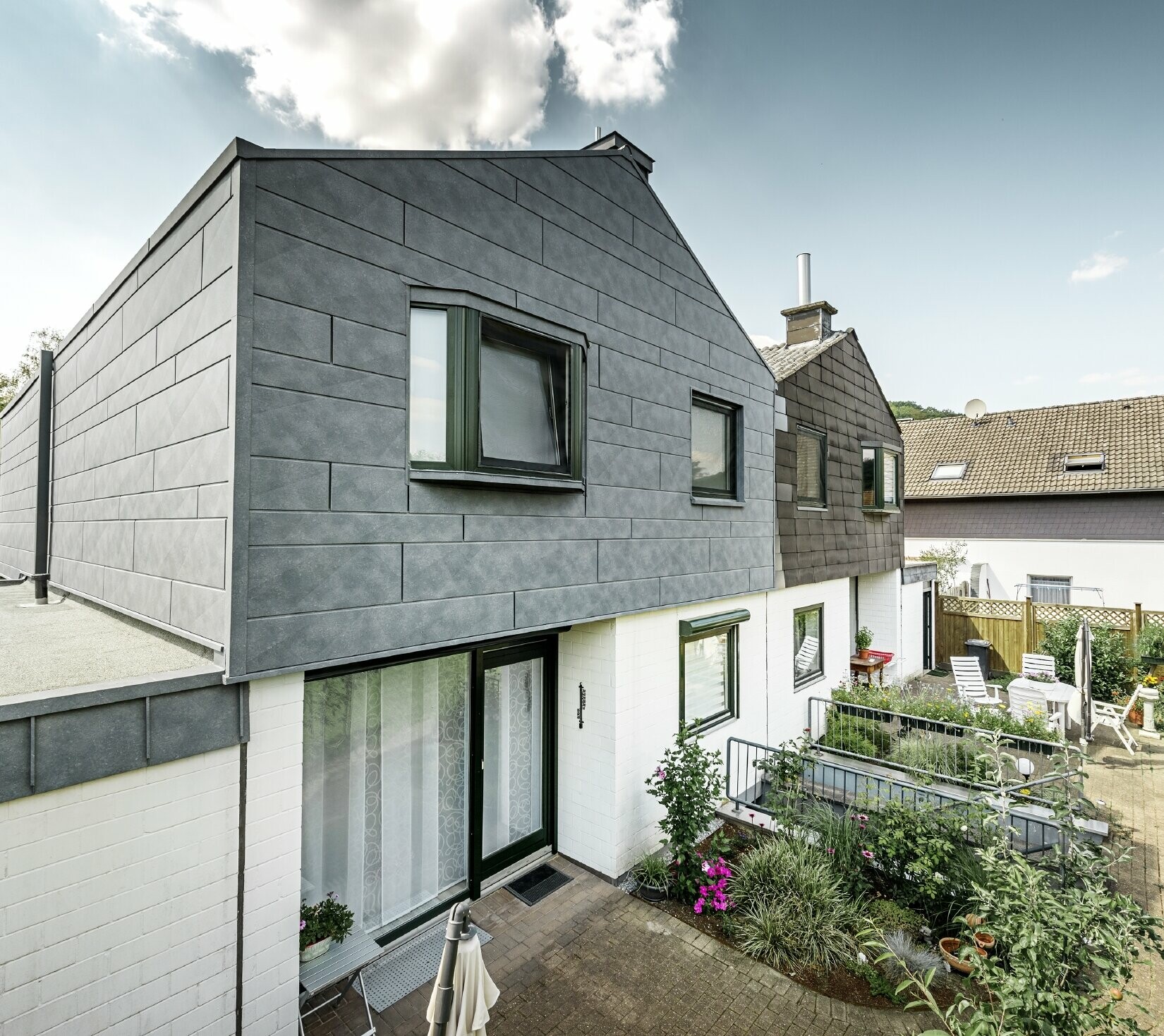 La partie haute du bâtiment de la moitié de maison jumelée a été rénovée avec l’élément de façade Siding.X PREFA en P.10 gris pierre. Juste à côté pour la comparaison, la moitié de maison avant la rénovation.