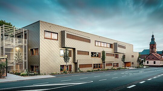 La photographie prise au niveau de la rue montre la nouvelle crèche d'Alzenau en dialogue dynamique avec le paysage urbain historique, notamment l'église remarquable en arrière-plan. La façade moderne, construite à partir de losanges de façade 44 x 44 couleur bronze, confère au bâtiment une élégance presque sculpturale. Des fenêtres judicieusement placées, des persiennes verticales en bois et des surfaces de façade décalées créent profondeur et rythme. À gauche, on aperçoit la cage d'escalier délicate avec sa structure métallique blanche, qui évoque une impression de légèreté moderne. La clarté du design, le choix de matériaux durables et la façade en aluminium de haute qualité font de ce bâtiment un projet phare d'intégration urbaine et de construction d'avenir grâce aux systèmes PREFA