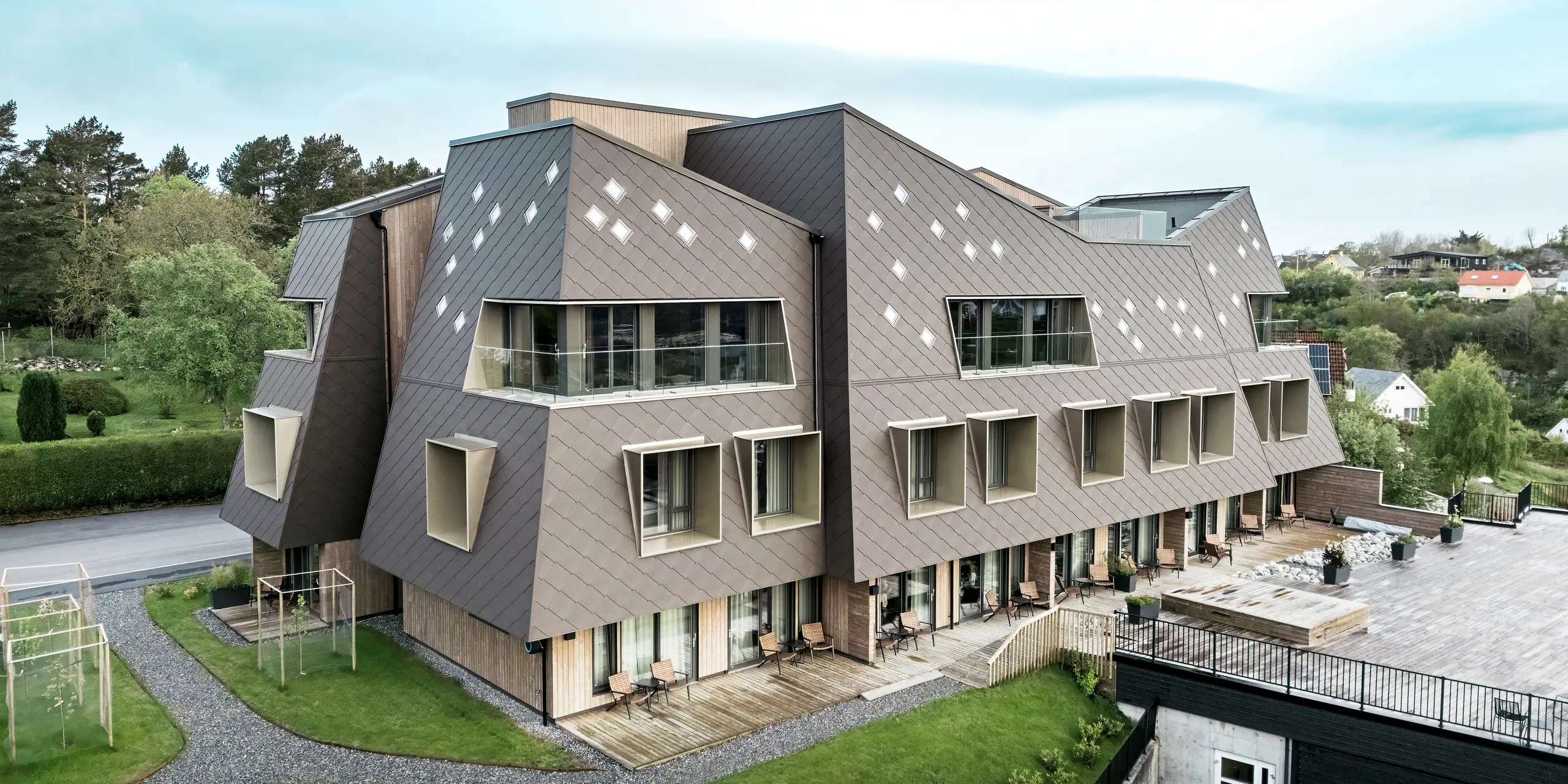 Vue extérieure de la maison Beckerwyc à Bekkjarvik, en Norvège, avec un accent sur les espaces extérieurs bien entretenus et la façade saisissante composée de losanges PREFA 44 × 44 en brun noisette. L'architecture sculpturale du bâtiment, combinée à une terrasse et des balcons spacieux, crée une connexion harmonieuse entre les espaces intérieurs et extérieurs. Le design futuriste et les couleurs naturelles s'intègrent bien dans le paysage environnant.