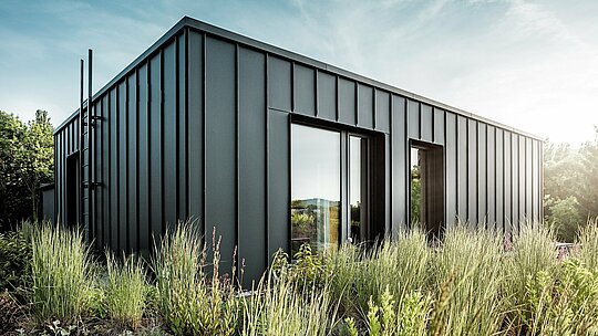 La photographie révèle la terrasse cubique et moderne d'une villa multigénérationnelle à Budapest, revêtue d'aluminium PREFALZ P.10 anthracite de haute qualité. Les joints debout, d'une structure nette, dessinent une élégante ligne verticale, particulièrement mise en valeur par la douce lumière du soleil. Entourée d'une végétation luxuriante, la terrasse offre un contraste saisissant entre l'architecture métallique moderne et la verdure environnante. La robustesse et la résistance aux intempéries des produits en aluminium PREFA témoignent d'une qualité d'exécution irréprochable et font de cette terrasse un élément architectural remarquable.