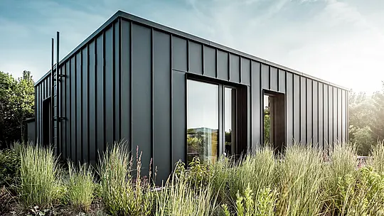 La photographie révèle la terrasse cubique et moderne d'une villa multigénérationnelle à Budapest, revêtue d'aluminium PREFALZ P.10 anthracite de haute qualité. Les joints debout, d'une structure nette, dessinent une élégante ligne verticale, particulièrement mise en valeur par la douce lumière du soleil. Entourée d'une végétation luxuriante, la terrasse offre un contraste saisissant entre l'architecture métallique moderne et la verdure environnante. La robustesse et la résistance aux intempéries des produits en aluminium PREFA témoignent d'une qualité d'exécution irréprochable et font de cette terrasse un élément architectural remarquable.