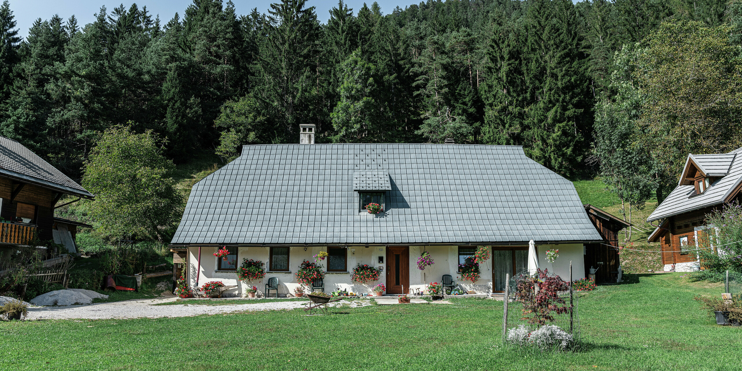 Une maison familiale pittoresque à Tarvisio, en Italie, est recouverte de tuiles PREFA robustes depuis 1972 : le grand toit en croupe avec revêtement en aluminium protège de manière fiable le bâtiment des intempéries alpines depuis des décennies. La façade est décorée de jardinières colorées et le jardin bien entretenu avec un coin salon et une petite cheminée vous invite à vous attarder. Entourée de conifères denses et de maisons traditionnelles en bois, la maison s'intègre harmonieusement dans le paysage naturel alpin.