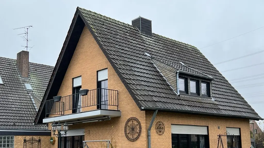 Maison individuelle en briques jaunes avec toit en pente raide et couverture de toit sombre avant la rénovation du toit ; côté jardin, avec balcon, lucarne, terrasse avec mobilier et pelouse au premier plan.
