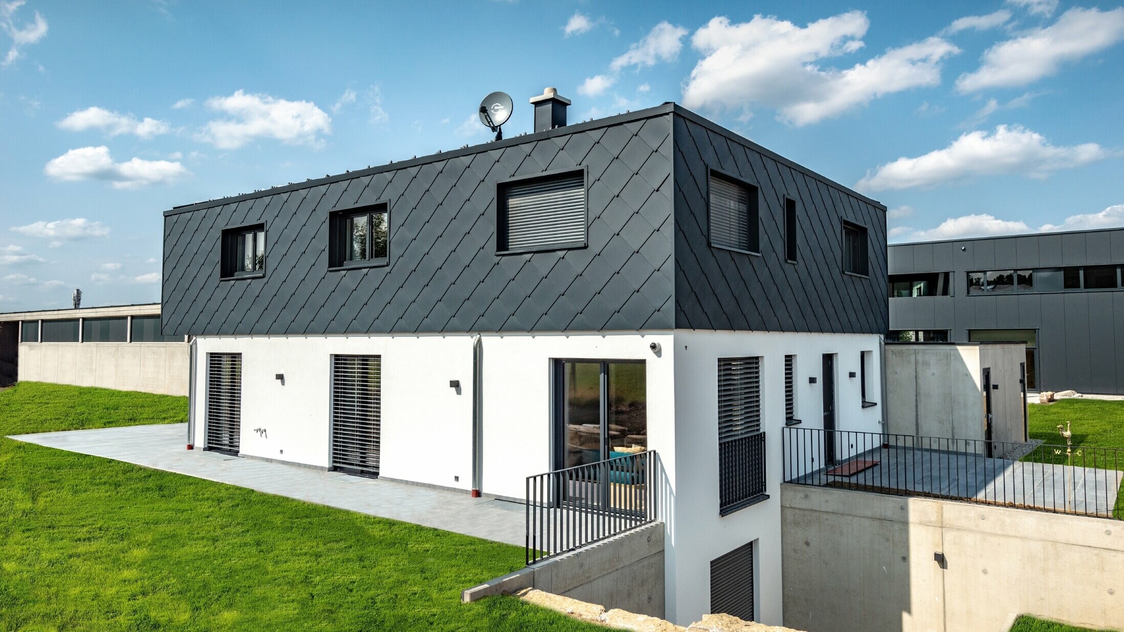 Nouvelle construction sobre à toit plat, le rez-de-chaussée est revêtu de crépi banc, l’étage supérieur est recouvert de grands losanges de façade PREFA couleur anthracite.