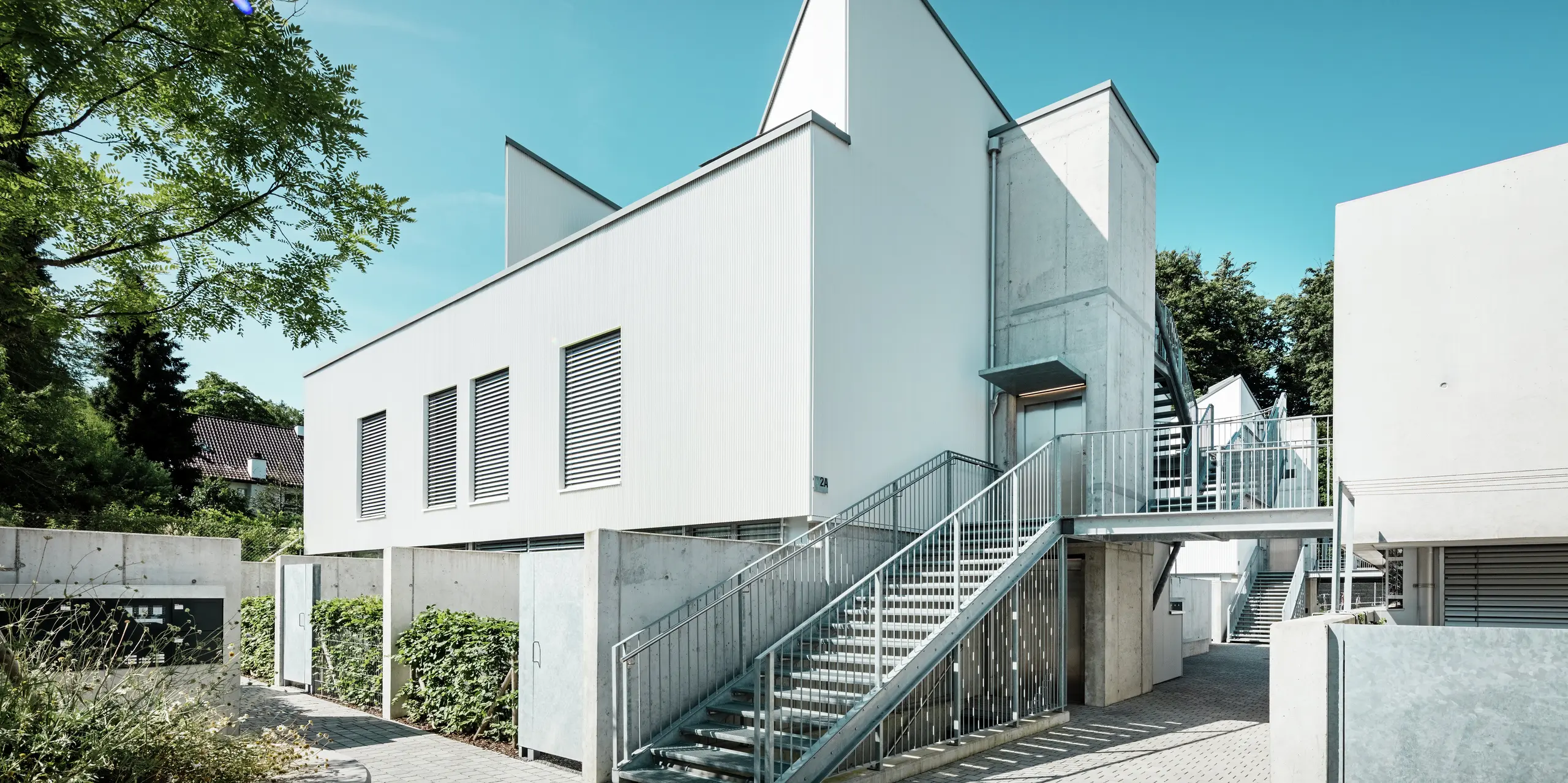 Un ensemble de mini-maisons modernes aux toits à deux pans remarquables, revêtues de profilés triangle PREFA P.10 Blanc Pur. La façade, simple et épurée, s'harmonise avec les éléments en béton apparent et la structure métallique environnante. Durable, la façade en aluminium PREFA assure une protection fiable contre les intempéries et garantit un aspect minimaliste et esthétique, nécessitant peu d'entretien.