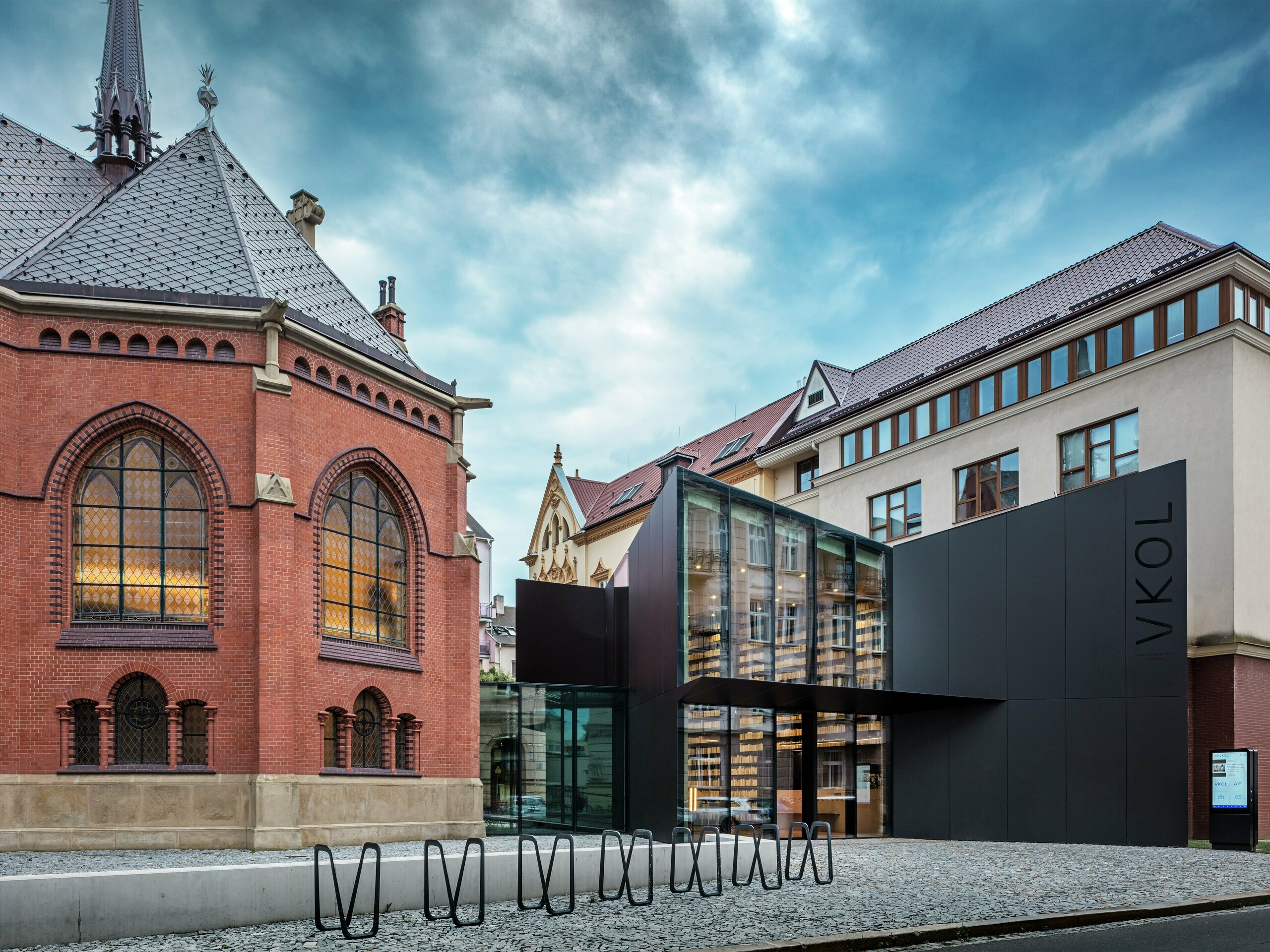 L'image illustre une fusion fascinante d'architecture historique et contemporaine à Olomouc. À gauche se dresse la majestueuse Église Rouge, avec ses fenêtres gothiques caractéristiques et ses murs de briques rouges, tandis que le nouveau bâtiment, à droite, contraste avec sa façade moderne en panneaux composites en aluminium noir PREFABOND. La façade vitrée du nouveau bâtiment laisse entrevoir la conception novatrice de la bibliothèque et confère à l'ensemble un caractère élégant et ouvert. La précision de la fabrication des panneaux PREFABOND garantit une apparence unifiée qui s'intègre harmonieusement au contexte historique tout en affirmant une vision contemporaine. L'architecture de ce projet allie parfaitement tradition et innovation, les matériaux étant non seulement esthétiques, mais aussi durables et respectueux de l'environnement.