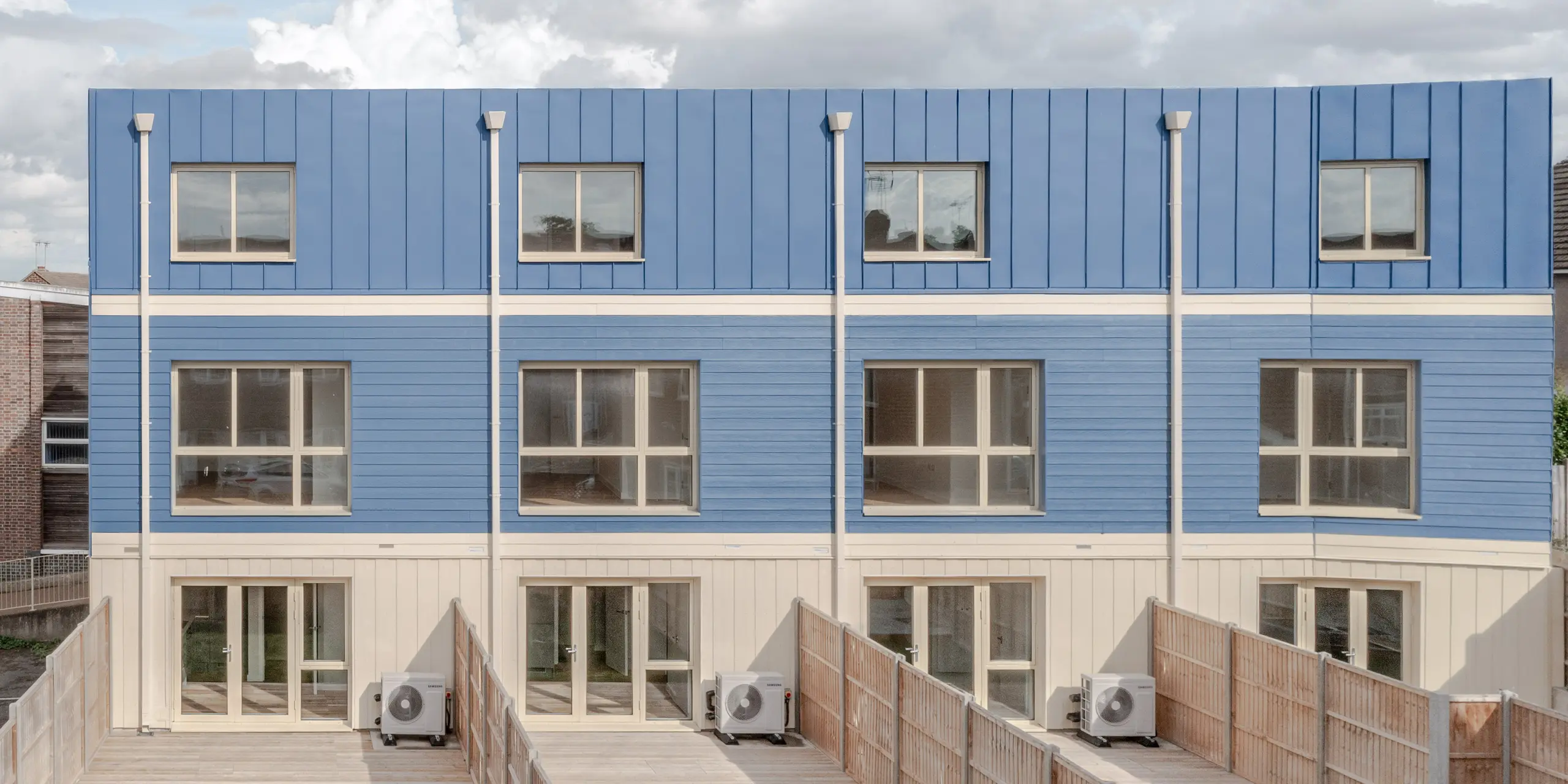 La façade côté jardin des maisons mitoyennes de Goldsmith Mews met en valeur le design saisissant de la façade et de la toiture PREFA FALZONAL bleu tourterelle, conférant aux bâtiments un caractère épuré et moderne. Les plis verticaux précis et les panneaux bleus créent une façade rythmée et dynamique. Les espaces lumineux du rez-de-chaussée et les larges fenêtres ouvrent l'architecture sur l'extérieur. L'aluminium PREFA de haute qualité offre une protection durable contre les intempéries, une excellente stabilité dimensionnelle et chromatique, et permet une conception durable et esthétique, à forte valeur ajoutée.