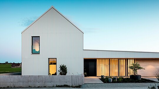 L'image présente une impressionnante maison individuelle à Freistadt, remarquable par son architecture géométrique audacieuse. La façade, réalisée en PREFALZ blanc pur aux plis précis, confère à la maison une élégance moderne. La structure verticale de la façade et le design épuré et minimaliste soulignent la qualité du matériau et garantissent une surface durable et résistante aux intempéries. Le toit à pignons abrupts, également revêtu de PREFALZ, protège des intempéries tout en contribuant à l'harmonie de l'ensemble. La clarté du design et l'utilisation de l'aluminium répondent parfaitement aux exigences esthétiques et fonctionnelles. Les fenêtres baignées de lumière et le portail d'entrée ouvert créent une atmosphère accueillante et offrent un contraste saisissant avec la modernité de la façade. Le choix du PREFALZ témoigne de l'ambition architecturale et représente une solution durable et nécessitant peu d'entretien pour les projets de construction pérennes.