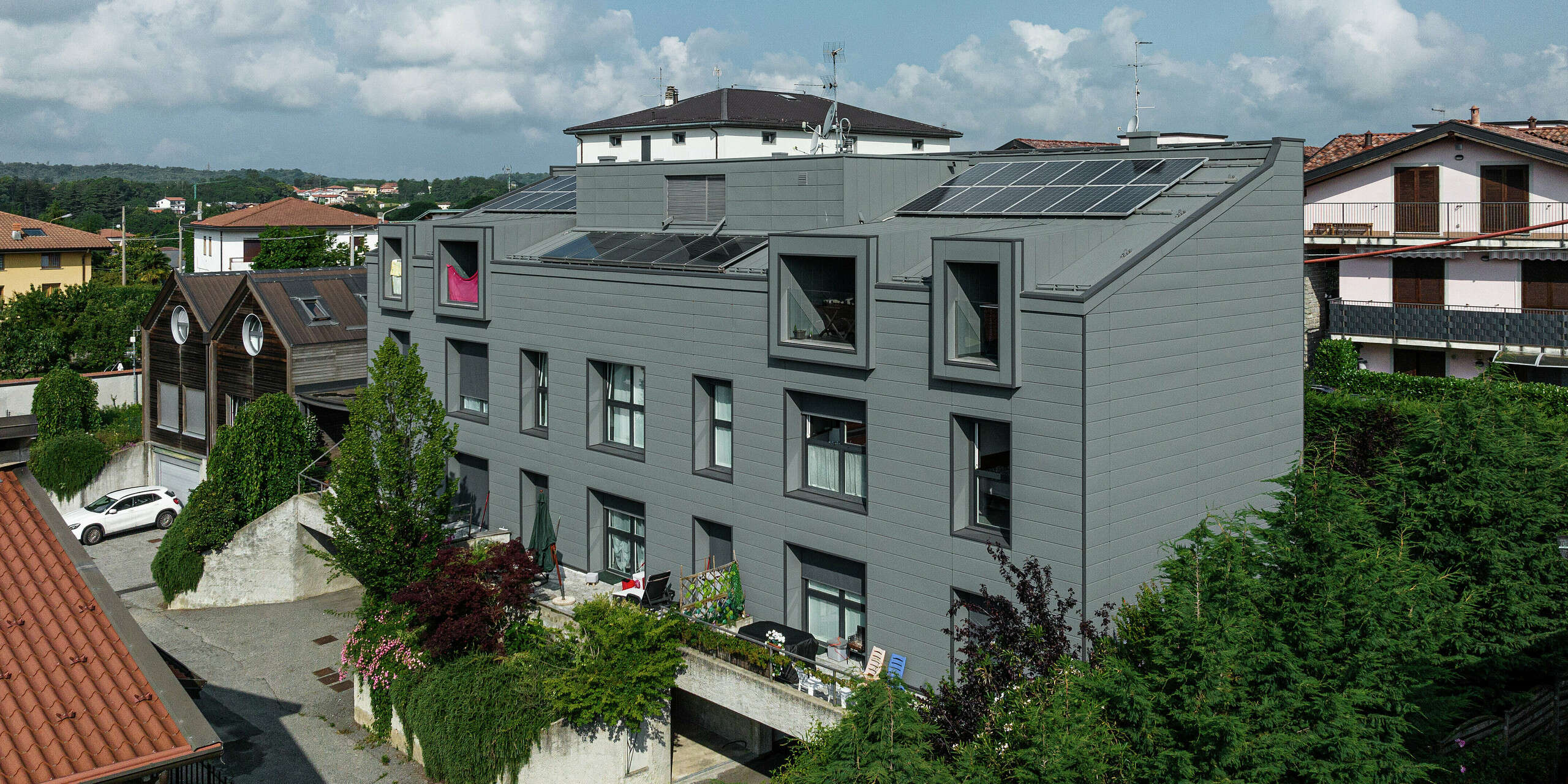 Vue panoramique du complexe résidentiel moderne de Faloppio, en Italie, avec une façade remarquable en bardages PREFA et un toit économe en énergie avec des modules photovoltaïques. Cette combinaison d’architecture innovante et de production d’énergie durable est un excellent exemple de construction moderne et durable. La façade en tôle assure une longévité et un entretien minimal, tandis que les cellules solaires fournissent une source d'énergie respectueuse de l'environnement.