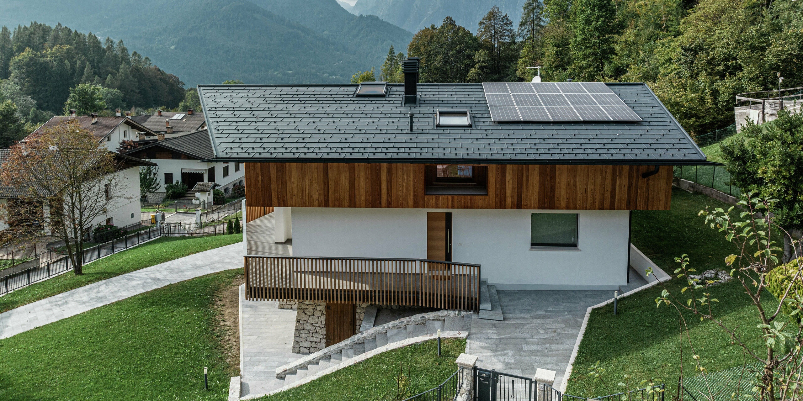 Vue latérale d'une maison individuelle moderne à Agordo, en Italie, avec des PREFA R.16 de couleur P.10 anthracite. Le toit en aluminium durable offre une protection optimale contre les intempéries et est idéal pour l'emplacement alpin. Un système de gouttière PREFA avec gouttière et tuyau de descente assure un drainage efficace de l'eau. La combinaison harmonieuse de la façade en bois, de la façade en plâtre blanc et du système photovoltaïque durable sur le toit souligne le design intemporel du bâtiment. Au premier plan, vous pouvez voir la zone d'entrée de la maison. En arrière-plan, vous pouvez voir un ciel nuageux et de nombreux arbres.
