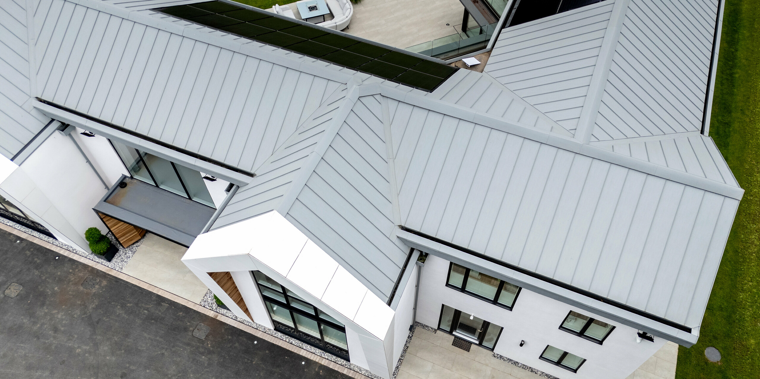 La vue aérienne révèle l'élégante toiture PREFALZ gris quartz de cette villa moderne située à Draycott, dans le quartier de Clay. La construction à joints debout confère à la toiture métallique une allure épurée et contemporaine, et elle s'intègre harmonieusement à son environnement. Équipée de modules photovoltaïques, la toiture souligne l'engagement écologique de la maison. La symétrie des éléments et la précision de la réalisation créent une impression d'ensemble esthétique, alliant fonctionnalité et design.