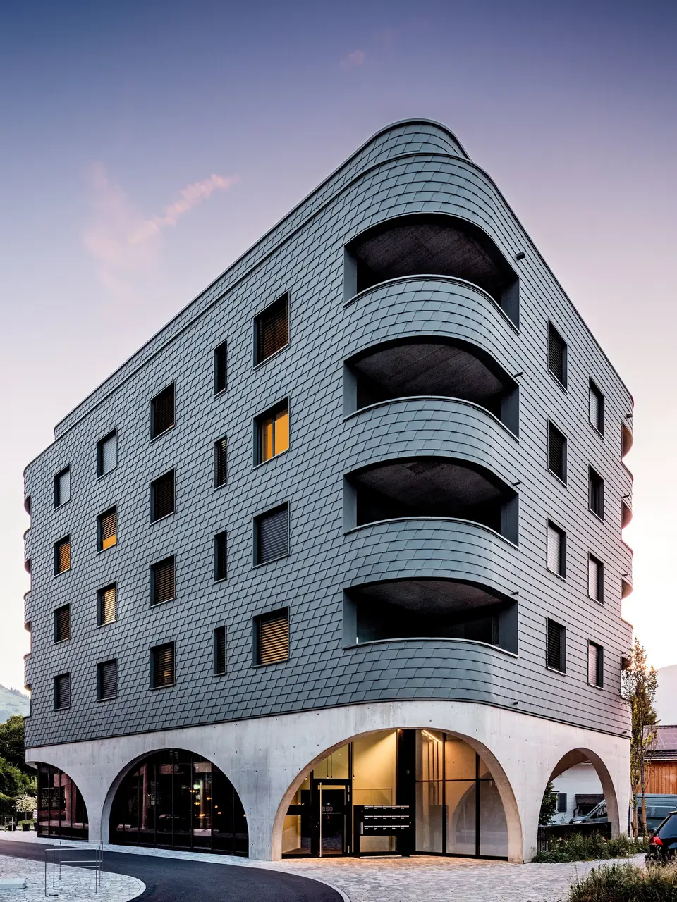 Un immeuble moderne à usage mixte à Buchs, à la forme triangulaire arrondie saisissante et à la façade continue en bardage PREFA gris souris P.10. L'enveloppe en aluminium, aux allures d'écailles, épouse la structure du bâtiment comme un reptile, soulignant ses courbes fluides et ses balcons profondément encastrés. Au-dessus d'un socle massif aux élégantes arcades en béton, la façade PREFA, légère et résistante aux intempéries, se distingue par son expressivité. La pose précise des bardeaux d'aluminium garantit une enveloppe homogène, durable et nécessitant peu d'entretien, tout en assurant une forte identité visuelle.