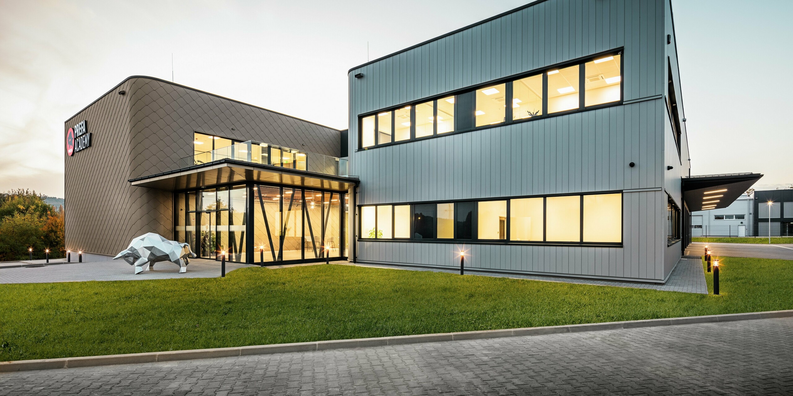 Le nouveau site PREFA à Törökbálint (HU) brille par son design moderne au crépuscule. La façade combine des losanges de façade PREFA 44 × 44 en bronze avec des Sidings PREFA en argent métallisé pour créer une enveloppe de bâtiment durable et résistante aux intempéries. De généreuses fenêtres créent une atmosphère lumineuse et conviviale, et la sculpture emblématique du taureau symbolise la force de la marque PREFA.