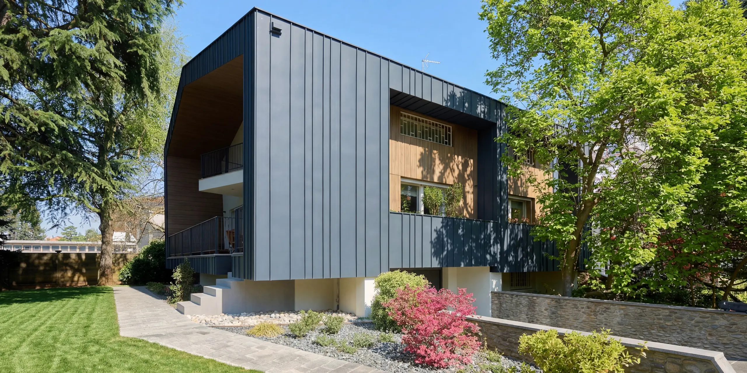 Vue de la Villa Castellanza et de sa remarquable façade PREFALZ couleur Anthracite P.10. Les lignes épurées des joints debout encadrent des éléments en bois chaleureux, conférant au bâtiment une allure moderne et élégante. Entourée d'une végétation luxuriante, la robustesse du bardage en aluminium PREFA est mise en valeur, alliant harmonieusement design, résistance aux intempéries et durabilité.