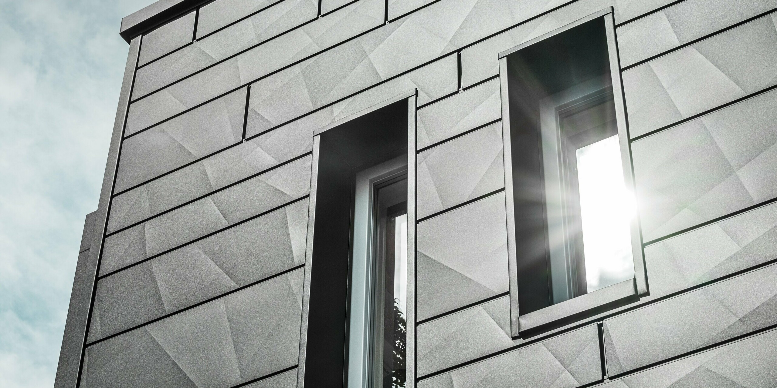 La façade moderne de la maison individuelle rénovée de Teuschnitz est ornée de panneaux géométriques Siding.X  PREFA en P.10 noir. Les encadrements de fenêtres en panneaux composites en aluminium noir s'intègrent harmonieusement à l'ensemble. Un rayon de soleil traverse l'une des fenêtres, soulignant la surface claire et texturée des panneaux de façade. L'esthétique de la façade est sublimée par le jeu subtil de la lumière et des matériaux.