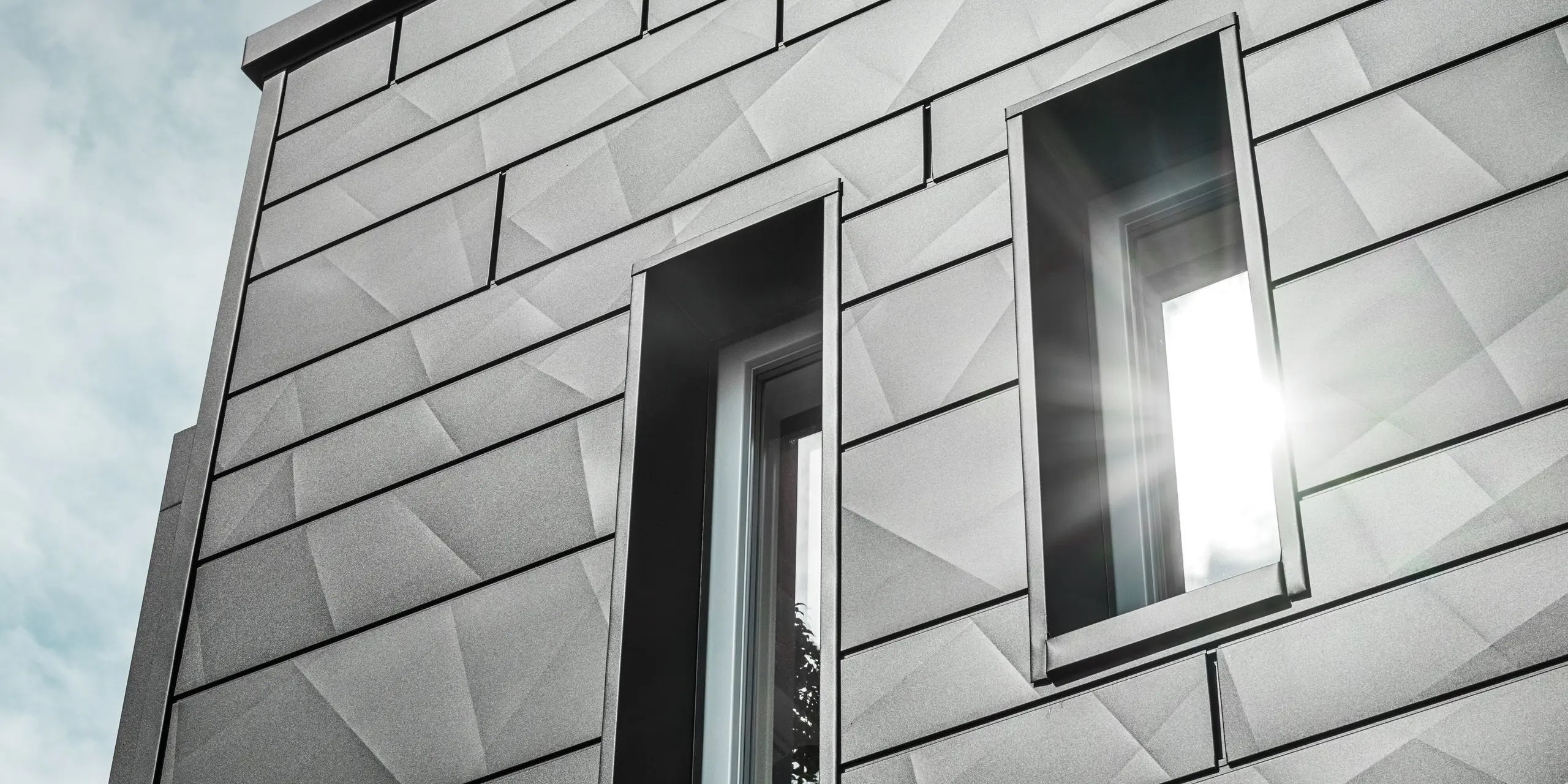 La façade moderne de la maison individuelle rénovée de Teuschnitz est ornée de panneaux géométriques Siding.X  PREFA en P.10 noir. Les encadrements de fenêtres en panneaux composites en aluminium noir s'intègrent harmonieusement à l'ensemble. Un rayon de soleil traverse l'une des fenêtres, soulignant la surface claire et texturée des panneaux de façade. L'esthétique de la façade est sublimée par le jeu subtil de la lumière et des matériaux.