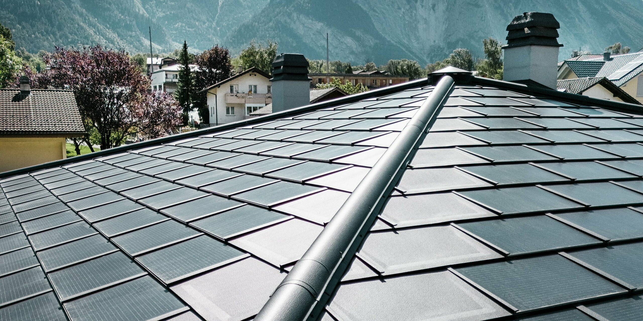 Les tuiles solaires innovantes PREFA de couleur P.10 noir s'intègrent parfaitement dans la conception du toit du domaine viticole Leukersonne à Susten. Le toit innovant en aluminium avec modules photovoltaïques intégrés génère une énergie solaire respectueuse de l'environnement sans aucun compromis visuel. Les R.16 PREFA ont été posés sur les bords. Au premier plan de l'image, on peut voir la crête du toit de la tente, réalisée avec précision. En arrière-plan, vous pouvez voir d'autres toits et le ciel bleu vif.