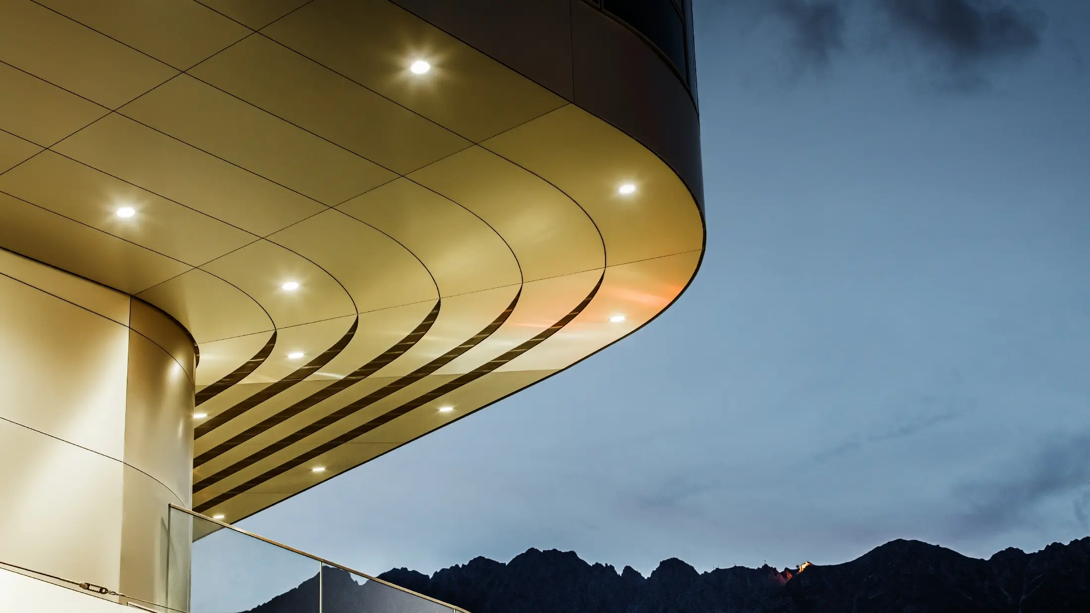 La photo montre un détail architectural impressionnant d'un panneau composite PREFA PEMA en aluminium brossé. Le panneau est légèrement bombé et présente des lignes fluides et élégantes qui sont particulièrement mises en valeur le soir grâce à un éclairage ciblé. La structure claire et l'aspect noble de l'aluminium du panneau complètent le design moderne de la façade et offrent une solution élégante et contemporaine pour une architecture de haute qualité.
