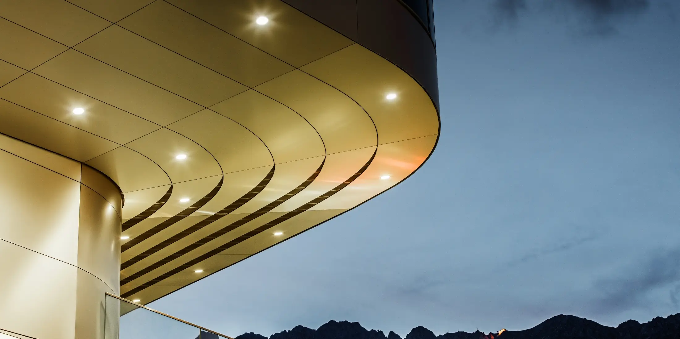 La photo montre un détail architectural impressionnant d'un panneau composite PREFA PEMA en aluminium brossé. Le panneau est légèrement bombé et présente des lignes fluides et élégantes qui sont particulièrement mises en valeur le soir grâce à un éclairage ciblé. La structure claire et l'aspect noble de l'aluminium du panneau complètent le design moderne de la façade et offrent une solution élégante et contemporaine pour une architecture de haute qualité.
