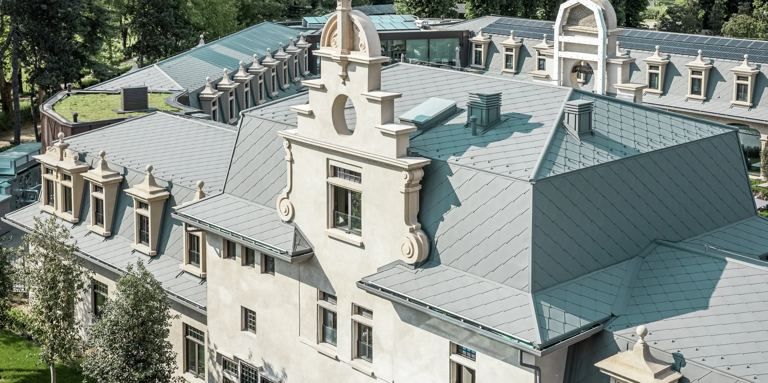 Vue détaillée du toit du parc thermal « De Montel – Terme Milano », avec sa toiture finement réalisée en losanges PREFA 44 × 44 P.10 gris souris. La géométrie complexe du toit, avec ses nombreuses lucarnes, corniches, pignons et éléments de ventilation, est magistralement exécutée. L'architecture historique est harmonieusement complétée par des éléments modernes, tels qu'une toiture plate végétalisée et le système photovoltaïque parfaitement intégré en arrière-plan.