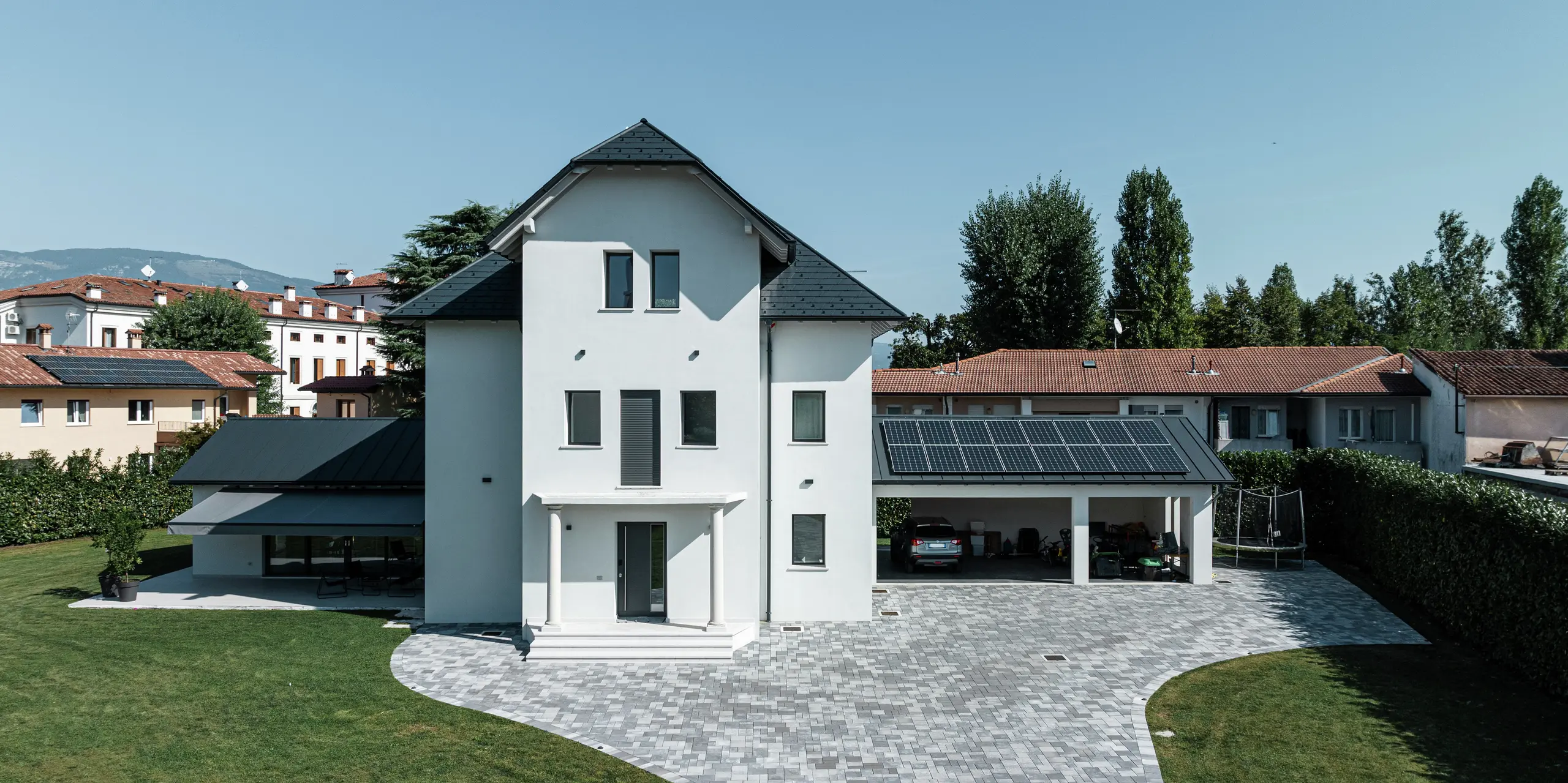 Vue de face d'une maison familiale moderne à Marano Vicentino, en Italie, avec des bardeaux de toiture PREFA durables P.10 Anthracite. Les dépendances adjacentes à gauche et à droite sont couvertes d'une toiture en aluminium à joint debout éprouvée - également en anthracite. Un système photovoltaïque au-dessus du garage fournit de l'électricité à partir d'énergie renouvelable. La façade en plâtre clair et le toit sombre forment un contraste élégant sur la maison unifamiliale de trois étages.