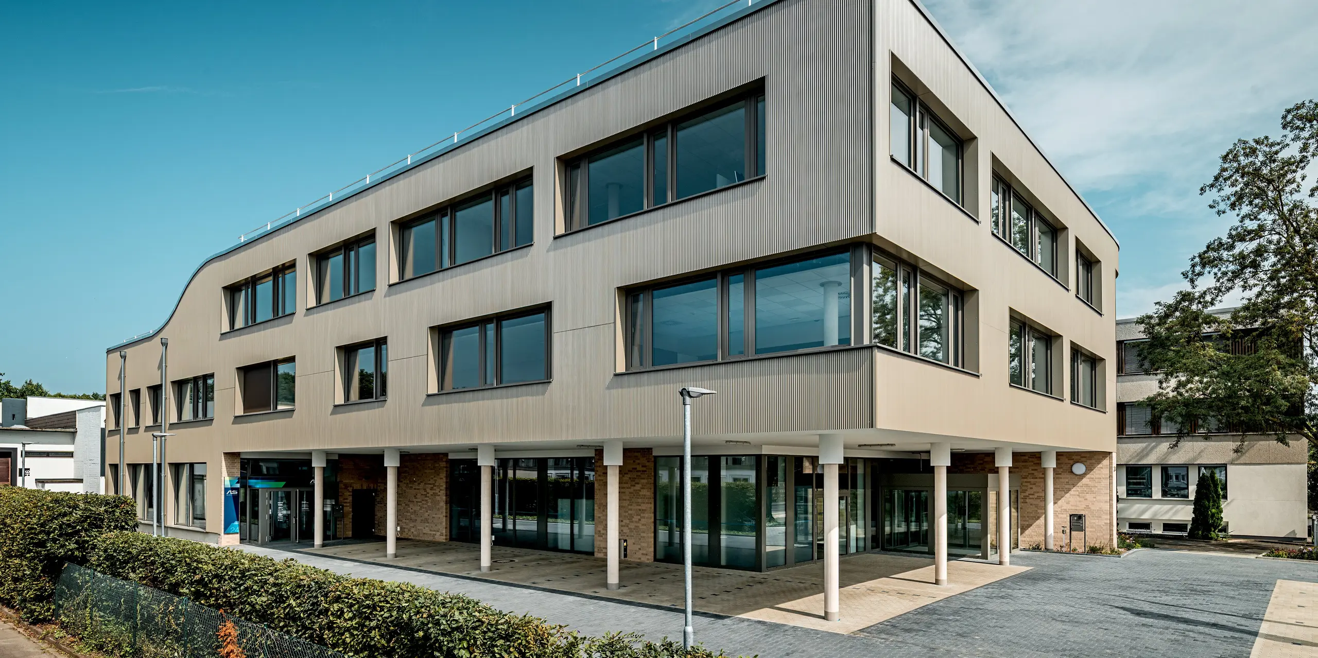 L'image présente l'entrée du bâtiment moderne Festo à Langenhagen, caractérisé par une façade incurvée remarquable. Cette façade est revêtue de profils extrudés triangle PREFA, créant une texture et une profondeur intéressantes. La pureté des surfaces vitrées et l'élégance des panneaux composites en aluminium assurent une harmonieuse combinaison de fonctionnalité et de design. Le bord supérieur légèrement arrondi du bâtiment est particulièrement remarquable et confère à l'ensemble une allure moderne et fluide.