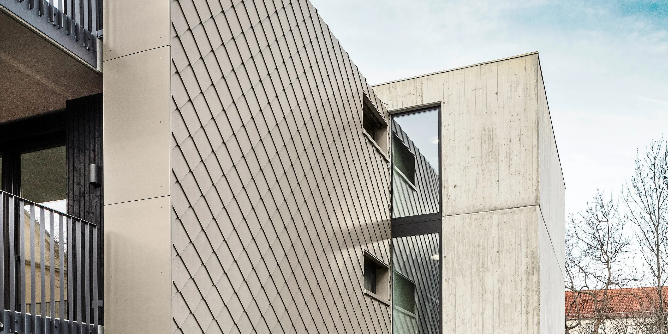 Zone d'accès à un immeuble d'appartements situé dans la Chemnitzer Straße, dans le quartier de Plauen, à Dresde. Le bâtiment est protégé par un système de toiture et de façade robuste PREFA en aluminium. Les losanges de toiture et de façade PREFA de haute qualité 29 × 29 en bronze assurent un aspect élégant et une protection durable. La photo montre la zone d'entrée, flanquée d'une élégante structure en losange et d'un mur en béton. La conception architecturale combine des matériaux robustes avec des lignes épurées et des éléments modernes, donnant au bâtiment un aspect contemporain et élégant.