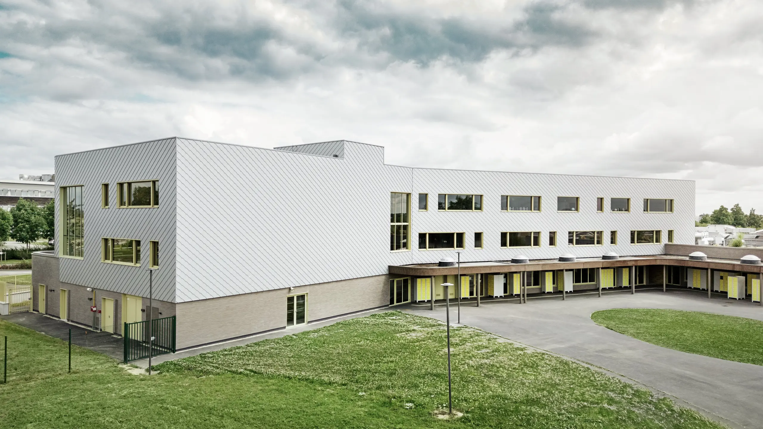 L'image montre l'arrière du collège Léontine Dolivet. Au premier plan, une vaste cour d'école pavée aux lignes épurées offre des espaces ouverts propices aux récréations et aux loisirs. Quelques espaces verts adoucissent l'environnement. À l'arrière-plan se dresse le bâtiment scolaire moderne, avec ses losanges de façade PREFA argent métallisé du plus bel effet, qui reflètent la lumière et créent un dynamisme certain. Plusieurs grandes fenêtres laissent entrer une abondante lumière naturelle. La scène a été photographiée en plein jour, sous un ciel légèrement nuageux, ce qui met particulièrement en valeur la surface métallique.