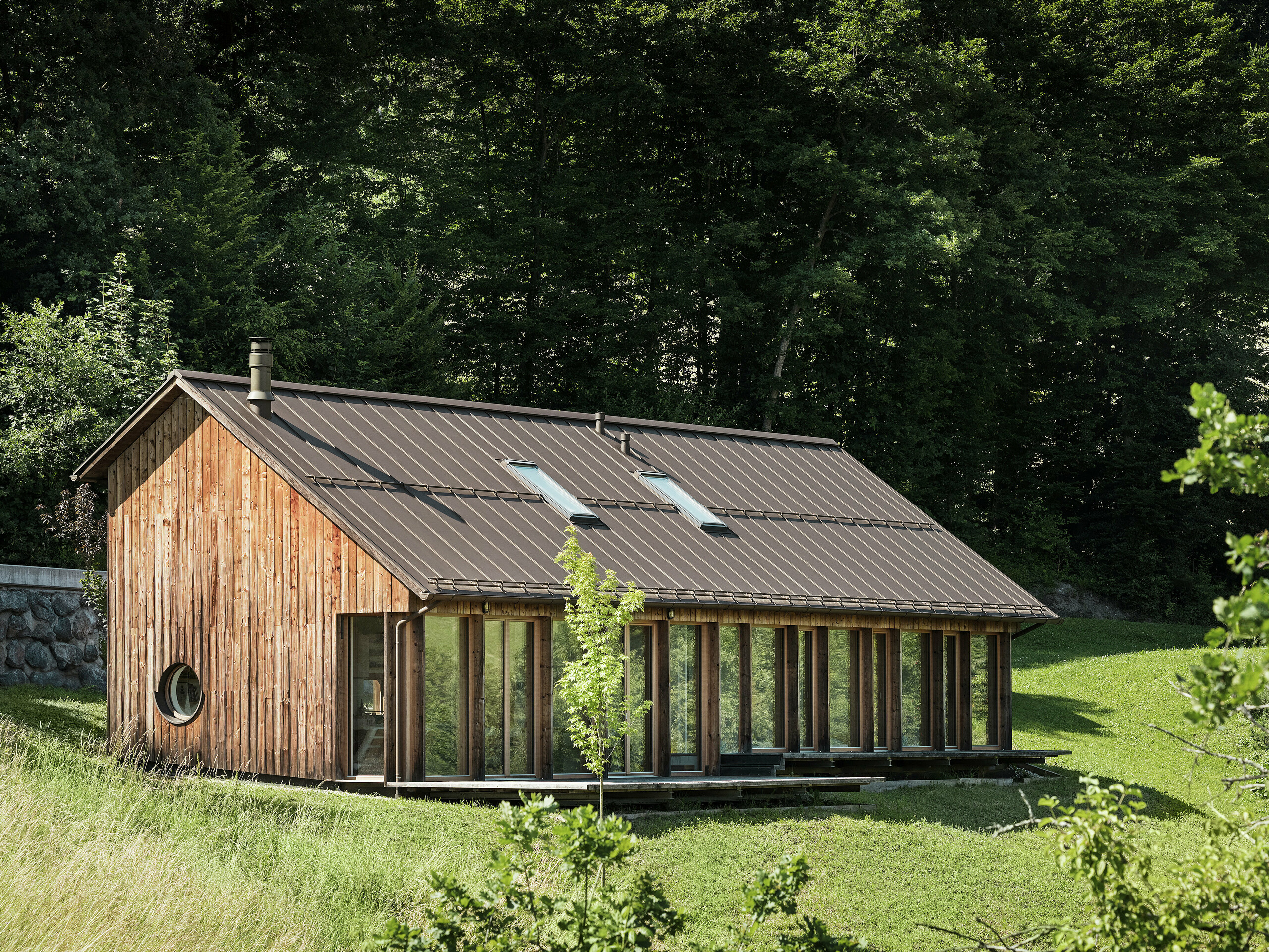 Maison de vacances en construction bois avec une grande façade vitrée et un toit PREFALZ de qualité en P.10 brun – harmonieusement intégrée dans le paysage vallonné verdoyant de Vinje, en Slovénie. Le toit en aluminium résistant séduit par sa durabilité, sa protection contre la corrosion et son esthétique moderne. Des fenêtres de toit intégrées assurent un éclairage naturel et une habitation écoénergétique. La maison en bois est entourée de nombreux arbres.