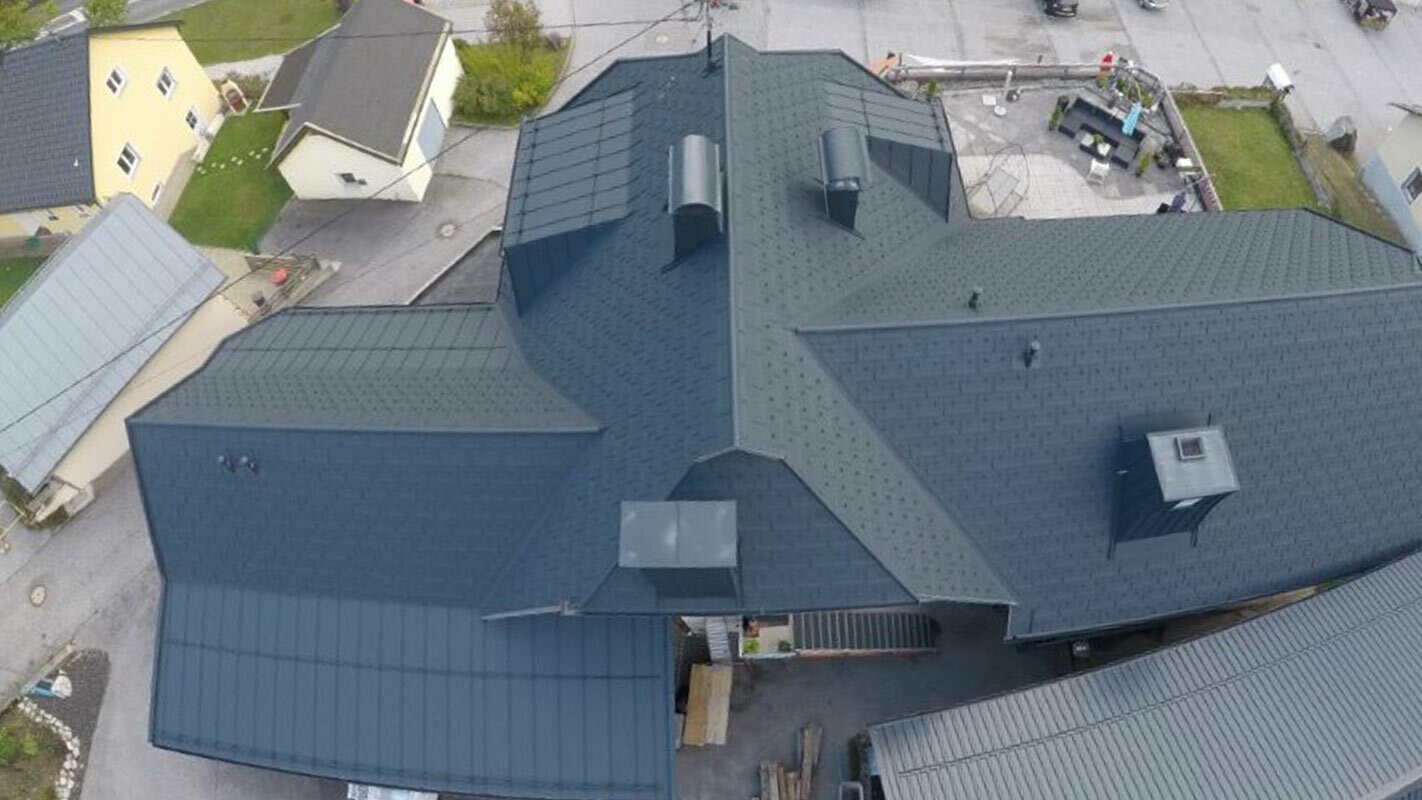 Rénovation d’une grande surface de toiture présentant de nombreux détails : noues, pignons et cheminées. Le toit est recouvert de tuiles R.16 en aluminium de PREFA, couleur anthracite.