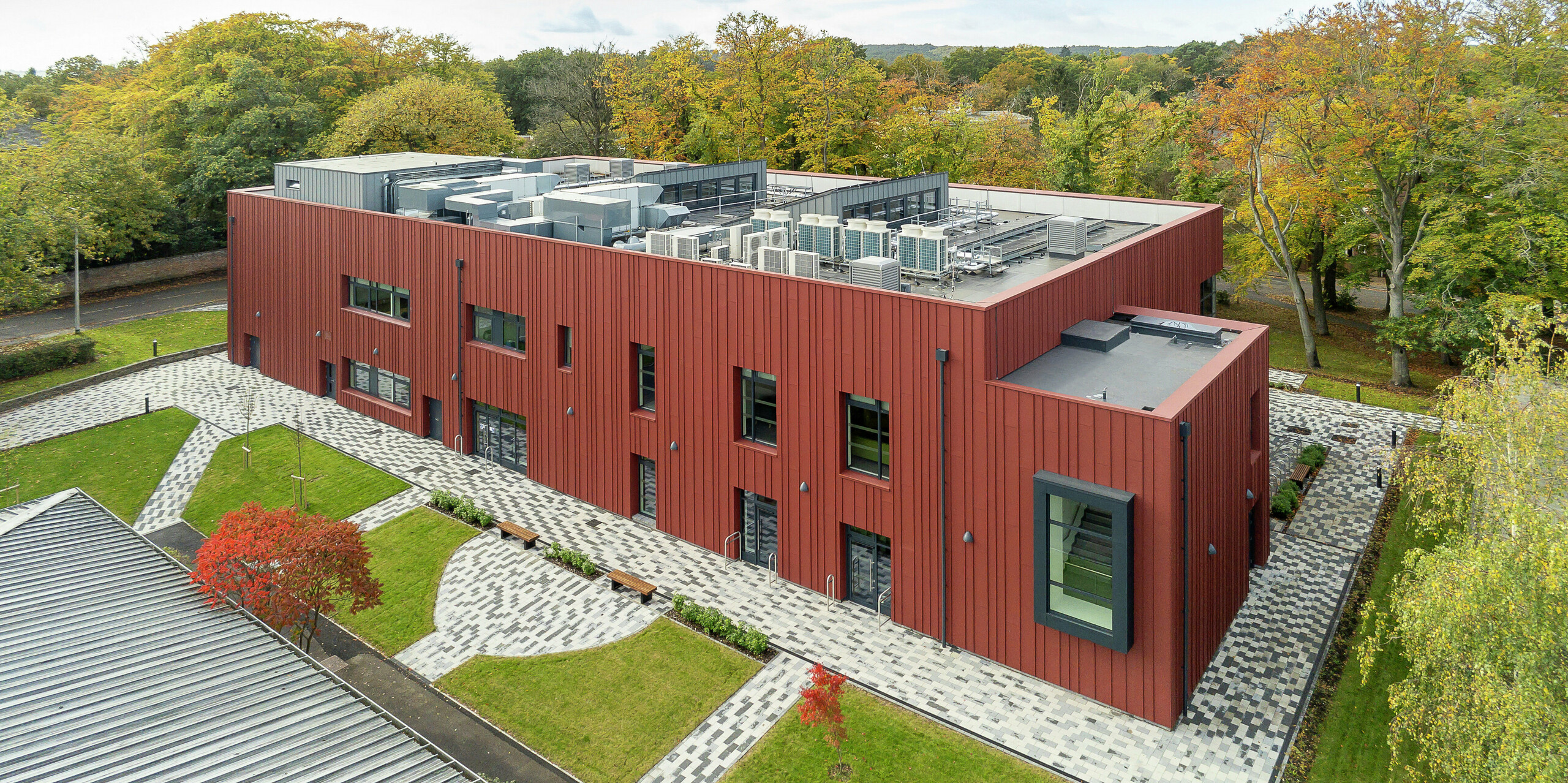 Une architecture en harmonie avec la nature : une vue plongeante sur le Soldiers' Centre de Pirbright. La façade à joint debout remarquable en PREFALZ en P.10 rouge oxyde s'intègre harmonieusement dans l'environnement verdoyant. Les cadres de fenêtres en porte-à-faux en P.10 anthracite apportent des accents modernes. L'espace extérieur structuré avec des chemins pavés, des espaces verts et des coins salon crée une atmosphère accueillante. Les installations techniques sont intégrées dans le toit plat, ce qui contribue à la technologie moderne du bâtiment.