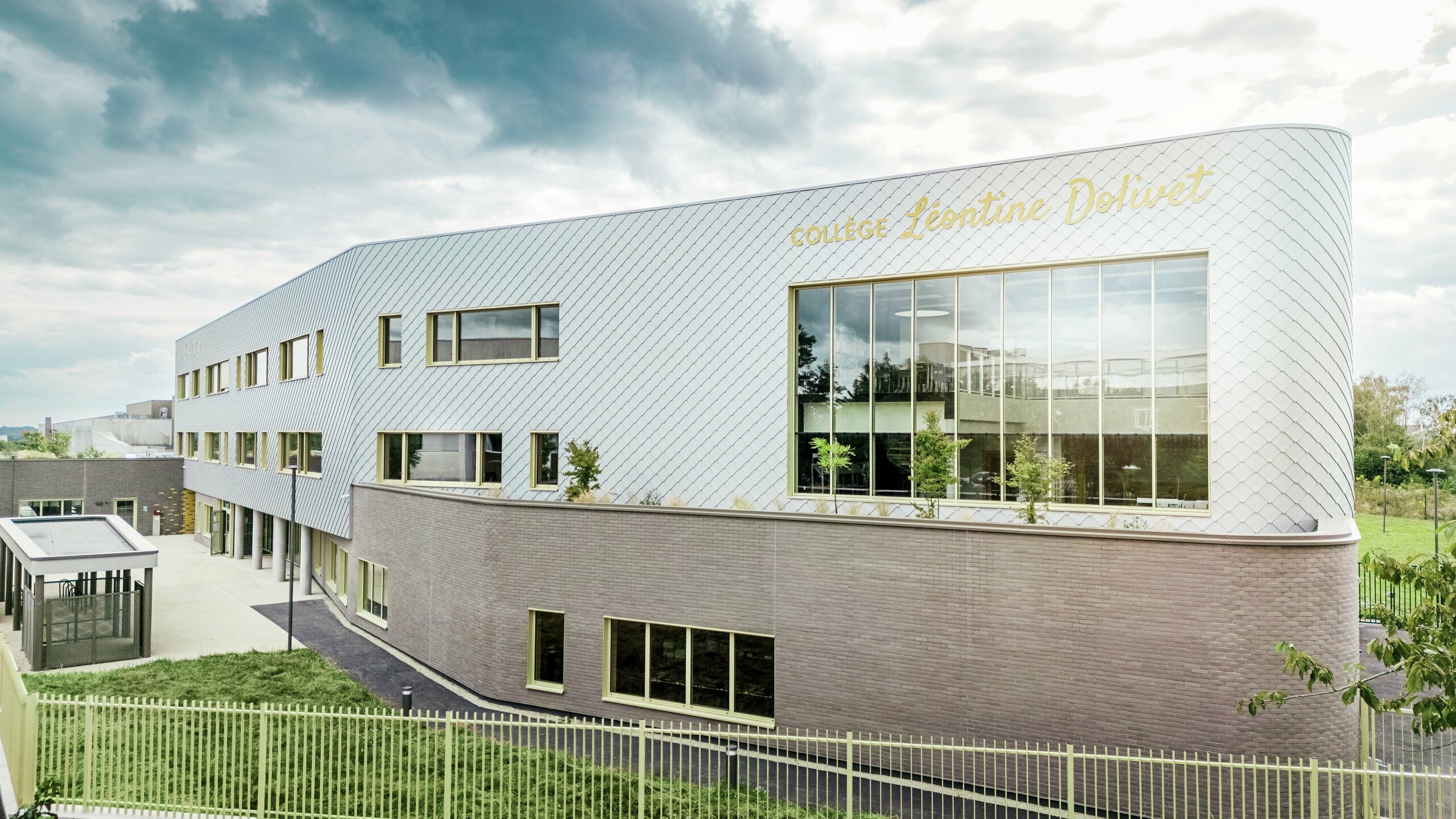 Vue latérale du Collège Léontine Dolivet à Cesson-Sévigné, dont la façade moderne en aluminium est composée de losanges PREFA argent métallisé. La structure diagonale crée un effet dynamique, tandis que les larges surfaces vitrées et les volets jaunes apportent des touches de couleur saisissantes. Au premier plan, des espaces verts et une clôture encadrent le bâtiment. Solution de façade durable, PREFA allie élégance, résistance aux intempéries et durabilité : une véritable prouesse architecturale aux lignes épurées et au métal scintillant.