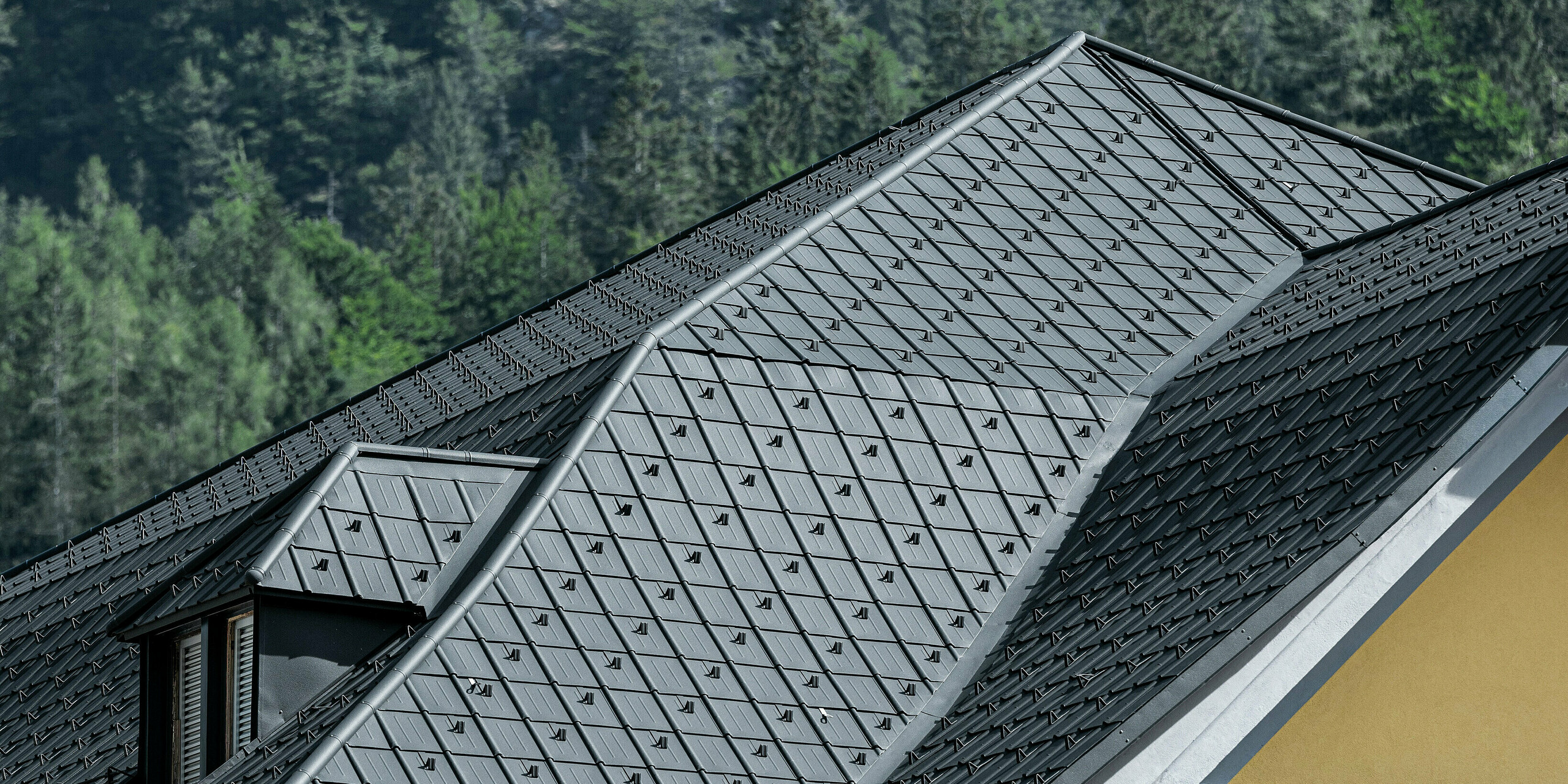 Vue détaillée de la toiture en aluminium PREFA de couleur anthracite (tuile P.10 anthracite) du Museo Cave del Predil. La finition de haute qualité, l'installation précise et les lucarnes intégrées sont clairement visibles. Idéalement adapté aux climats alpins et aux fortes charges de neige. On peut voir des pentes boisées de montagnes en arrière-plan.