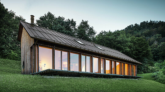 Ambiance du soir autour de la maison en bois moderne à Vinje, avec une façade vitrée continue et un toit en aluminium PREFALZ durable de couleur P.10 brun. La couverture de toit PREFA, résistante aux intempéries, offre une protection fiable et s’intègre parfaitement dans l’environnement naturel. Le design allie confort de vie, efficacité énergétique et durabilité esthétique.