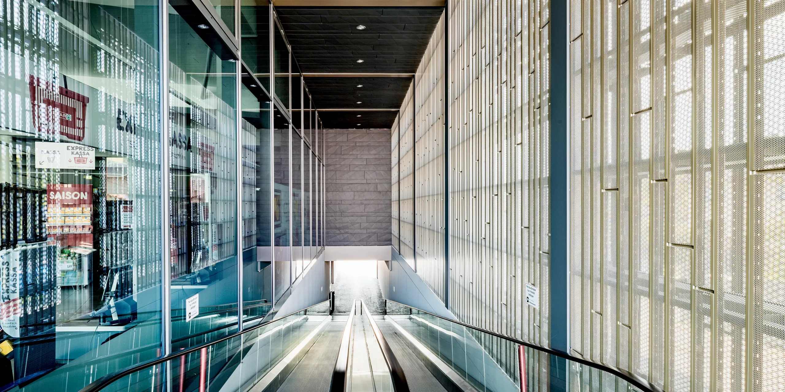 L'image révèle l'impressionnant hall d'entrée du centre commercial de Rohrbach, qui séduit par l'alliance de matériaux modernes et d'un design fonctionnel. Au premier plan, on aperçoit l'escalator menant aux niveaux inférieurs du bâtiment. Les murs sont revêtus de panneaux de bardage anthracite perforés, installés avec précision. Leur texture subtile et le jeu subtil de lumière qu'ils créent confèrent à l'espace une atmosphère agréable. Outre leur aspect esthétique, ces panneaux assurent une ventilation et une transmission optimales de la lumière naturelle, baignant ainsi l'intérieur de lumière naturelle. Les larges baies vitrées à gauche offrent une vue dégagée sur l'extérieur, renforçant l'impression d'ouverture et de transparence.