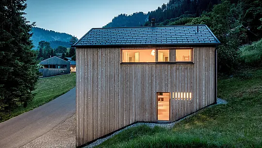 Face au spectaculaire paysage montagneux d’Ebnit, la maison Immi apparaît comme un chaleureux refuge au cœur de la forêt. Les fenêtres éclairées mettent en valeur la sobre façade en sapin blanc, tandis que le toit solaire PREFA noir souligne la silhouette épurée du bâtiment. Les tuiles solaires intégrés et les R.16 en P.10 noir produisent de l’énergie durablement tout en offrant une protection robuste et durable contre les intempéries en milieu alpin.
