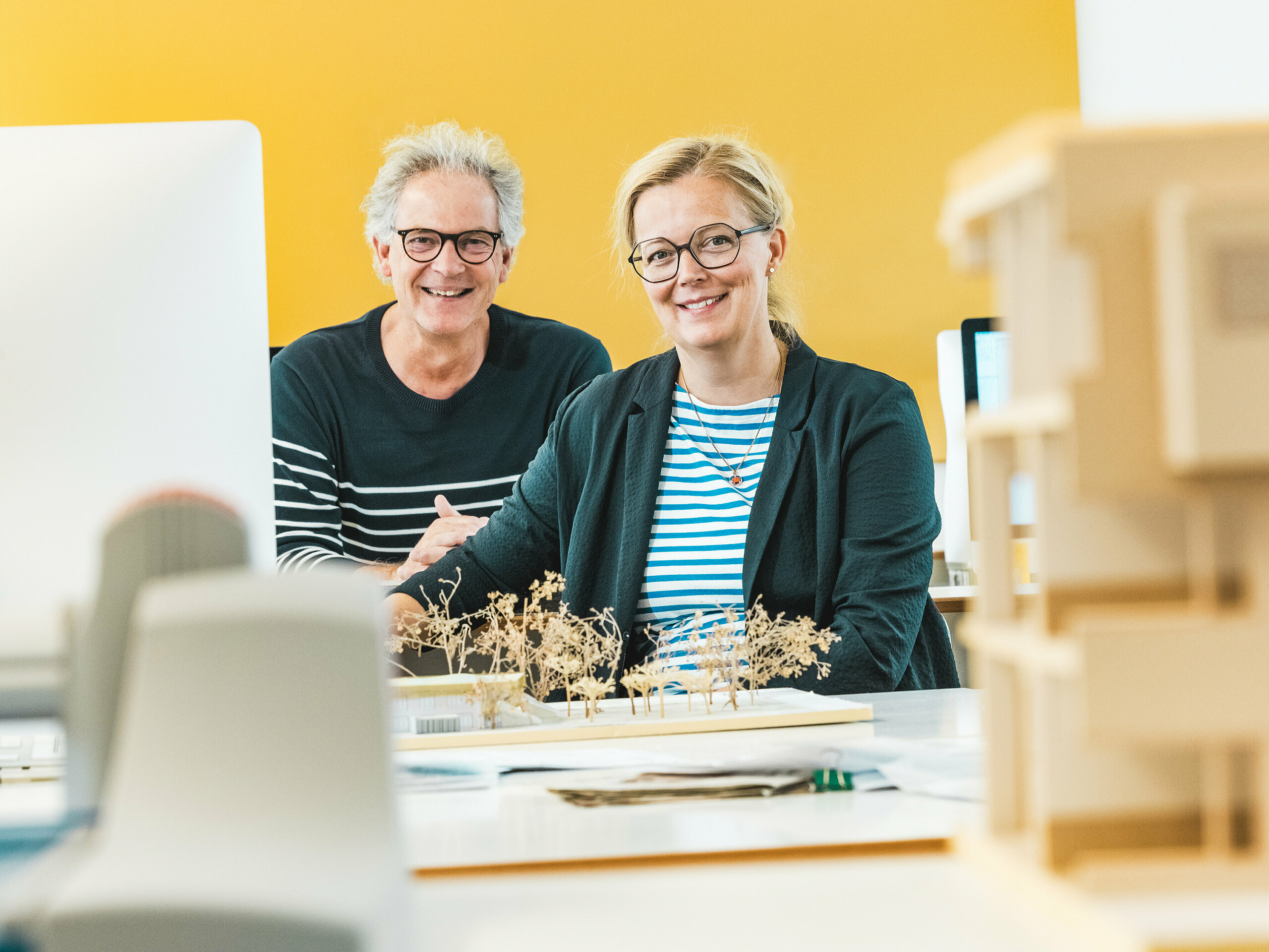 Les architectes Claudia et Theo Härtner, rayonnants, posent devant une maquette. Ce couple d'architectes conçoit la crèche d'Alzenau, un bâtiment hybride novateur doté d'une façade en aluminium PREFA. Au premier plan, des arbres miniatures symbolisent le concept global de développement durable. La crèche d'Alzenau allie une architecture innovante à un urbanisme responsable et met en valeur la polyvalence des losanges de façade 44x44 PREFA couleur bronze. Durable, facile d'entretien et 100 % recyclable, la façade en aluminium unifie visuellement les espaces résidentiels et la crèche en un ensemble harmonieux. Cette image capture l'énergie créative du processus de conception et souligne les atouts esthétiques et écologiques des produits PREFA.