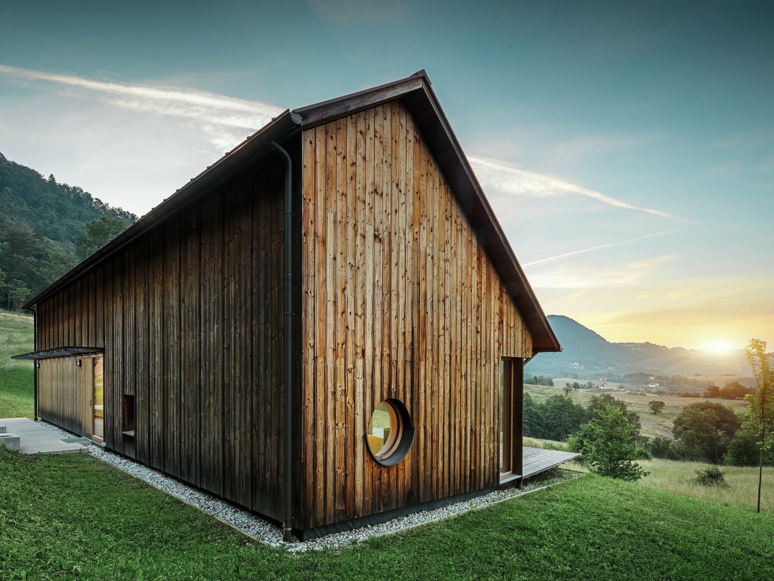Cette photo pleine d’atmosphère montre une maison en bois à Vinje au coucher du soleil, avec un toit en aluminium PREFALZ robuste de couleur P.10 brun. Le toit à joint debout de PREFA, résistant aux intempéries, est durable, nécessite peu d’entretien et s’intègre parfaitement dans l’environnement naturel. L’association de lignes épurées, d’une façade en bois et d’une ouverture de fenêtre arrondie souligne le minimalisme architectural. Le toit à deux pans, volontairement asymétrique, apporte une touche unique à l’ensemble.