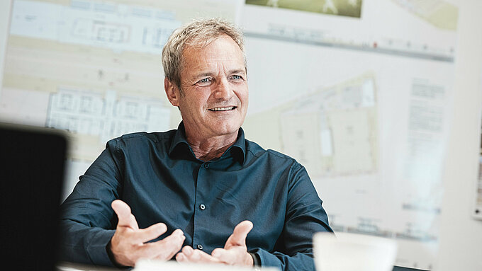 Portrait de l'architecte Max Stoisser, associé fondateur de planconsort, dans une conversation. Il porte une chemise bleu foncé et fait des gestes pendant qu'il parle. En arrière-plan, vous pouvez voir des plans et des visualisations du centre événementiel ZIB Nikolai.