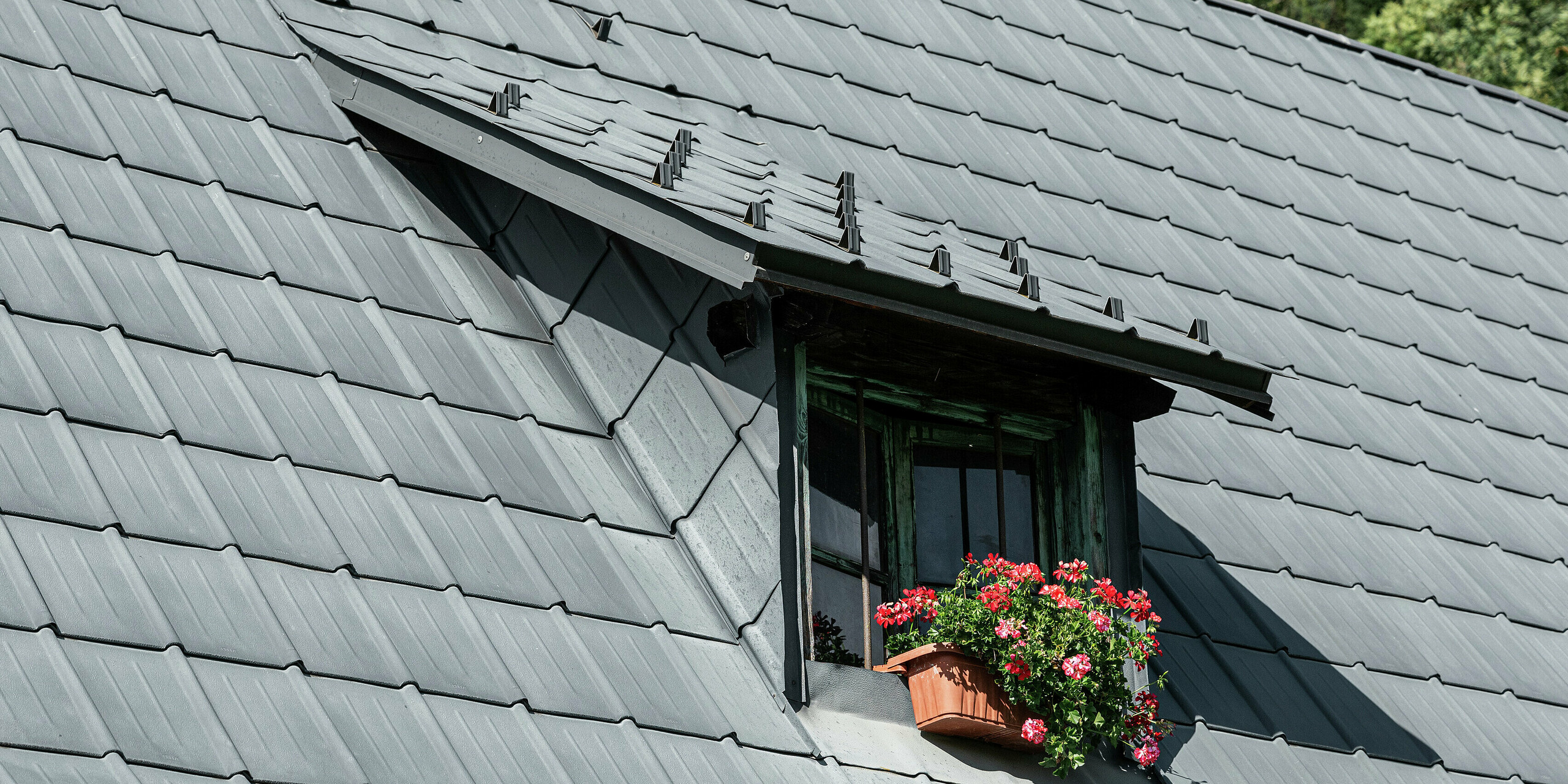 La maison familiale de Tarvisio, en Italie, recouverte de tuiles PREFA durables depuis 1972, impressionne par sa lucarne frappante au milieu du toit en croupe. Cette lucarne s'intègre harmonieusement dans la spacieuse toiture en aluminium et apporte une lumière supplémentaire dans le grenier. Les tuiles durables protègent la maison de manière fiable contre les conditions météorologiques alpines depuis des décennies. La lucarne décorée de fleurs apporte un accent attrayant et souligne la combinaison réussie de fonctionnalité et d'esthétique.