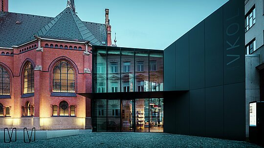 L'image illustre la fusion remarquable entre architecture historique et moderne à Olomouc. L'imposante église rouge, de style gothique, avec ses vitraux ornés et sa haute flèche, est mise en valeur par un nouveau bâtiment moderne à la façade noire minimaliste, réalisée en panneaux composites en aluminium PREFABOND. Les surfaces vitrées transparentes de ce nouveau bâtiment reflètent le paysage historique environnant et offrent un contraste saisissant avec la structure massive de l'église. La précision de la mise en œuvre des matériaux modernes, alliée à l'harmonieuse fusion de l'ancien et du nouveau, confère au bâtiment une élégance contemporaine tout en respectant l'histoire du lieu. Cette combinaison de matériaux innovants et durables comme PREFABOND garantit à la fois esthétique et fonctionnalité.