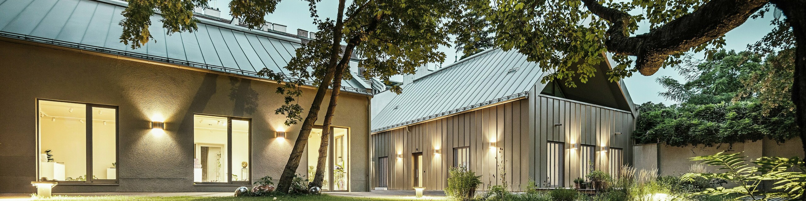 Vue nocturne du Studio Comploj à Vienne, entouré d'arbres et d'espaces verts. La façade et le toit en aluminium PREFALZ en P.10 bronze Prefa s'intègrent harmonieusement dans l'environnement.