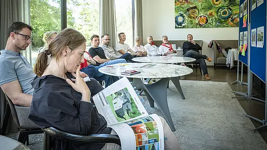 PREFARENZEN 2027 : Débat d’experts dans une salle de séminaire lumineuse : Paulina Frey (Directrice Marketing PREFA CH) présente un imposant ouvrage d’architecture illustrant des projets utilisant l’aluminium PREFA, tandis que les autres membres du jury écoutent attentivement. De larges baies vitrées laissent entrer la lumière naturelle et offrent une vue sur la verdure environnante. L’atmosphère est propice à une analyse approfondie, à des échanges constructifs et à une évaluation du design et de la fonctionnalité. Les produits en aluminium PREFA symbolisent des toitures et des façades durables et esthétiques, qui favorisent l’innovation, la qualité et l’inspiration dans les projets architecturaux.