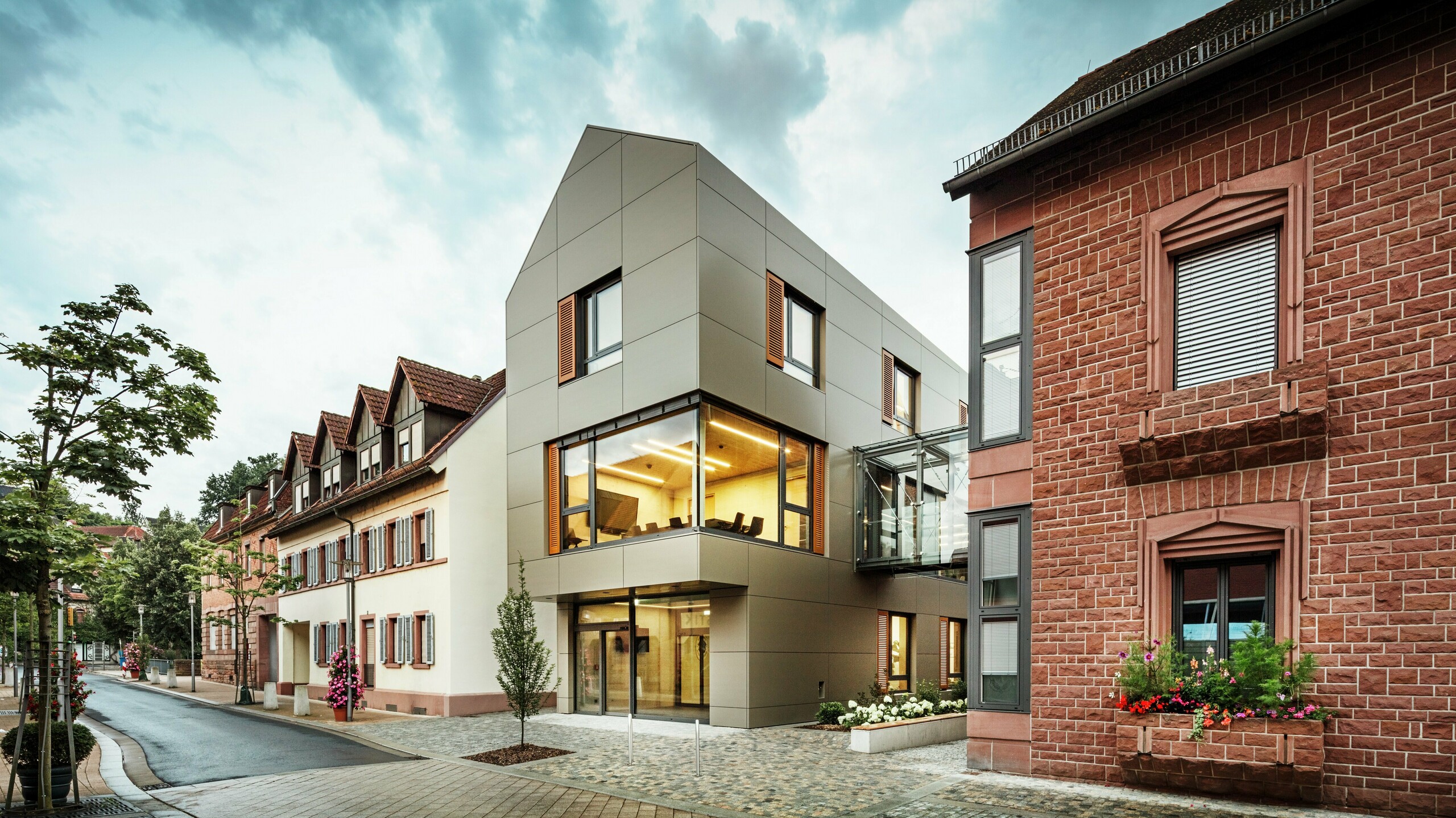 La photo montre la mairie moderne d'Alzenau, revêtue de panneaux composites en aluminium PREFA dans un ton bronze chaleureux. La façade est interrompue par de grandes surfaces vitrées qui offrent une vue sur l'intérieur, éclairé par une lumière chaude. La passerelle vitrée à droite de l'image relie le bâtiment à un bâtiment traditionnel en briques situé à proximité, créant ainsi un contraste intéressant entre l'architecture moderne et historique.